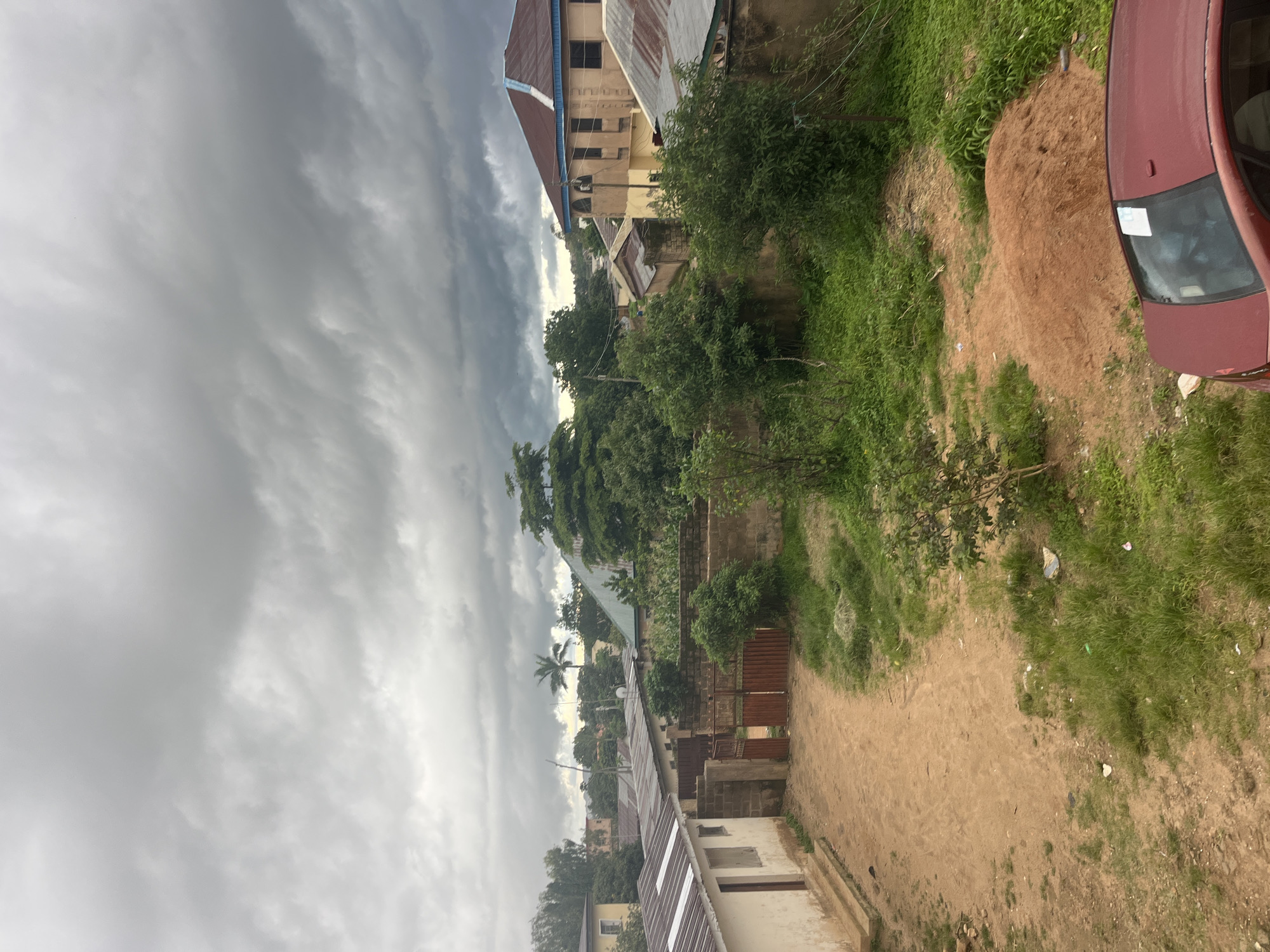 This is a medium-shot, eye-level photograph taken outdoors in Jos, Nigeria, under a cloudy sky. The scene depicts a dusty, unpaved compound with several buildings and dense vegetation. In the foreground, a portion of a maroon car is visible. The compound features a light-colored building with a dark roof on the left, and a red gate made of metal bars in the center, leading to a dirt path. Beyond the gate, more buildings with corrugated metal roofs are visible, along with a larger, tan-colored building with a blue-edged roof in the background to the right. Lush green trees and bushes are scattered throughout the scene, with some areas of overgrown grass and a pile of reddish-brown earth. The sky is dominated by heavy, dark gray clouds, suggesting an impending storm. There are no people visible in the frame.