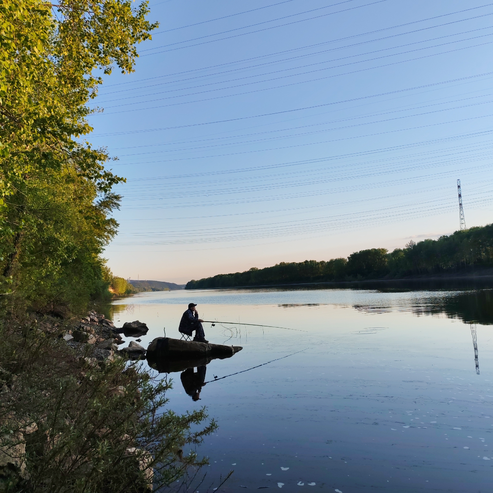 The image depicts a serene riverside scene, likely set in Chaikovsky, Russia, during the late afternoon or early evening. The clear, pale blue sky, gradually warming towards the horizon, suggests tranquil weather conditions.
The main subject is a person, dressed in dark clothing and a cap, seated on a small folding chair atop a large rock or log extending into the water. They are engaged in fishing, holding a long rod that stretches over the calm river surface. The person's reflection, along with the fishing rod, is clearly mirrored in the still water.
The foreground on the left features a rugged riverbank covered with rocks and dense green foliage. The river itself is wide and placid, reflecting the sky and the distant landscape. On the far bank, a continuous line of lush green trees forms a dense forest. In the distance on the right, a tall, metallic transmission tower stands prominently against the sky. Multiple parallel power lines crisscross the upper portion of the sky, indicating overhead utilities. The overall atmosphere is one of peaceful solitude and patience, as the angler enjoys the quiet beauty of the natural surroundings. There is no visible text in the image.