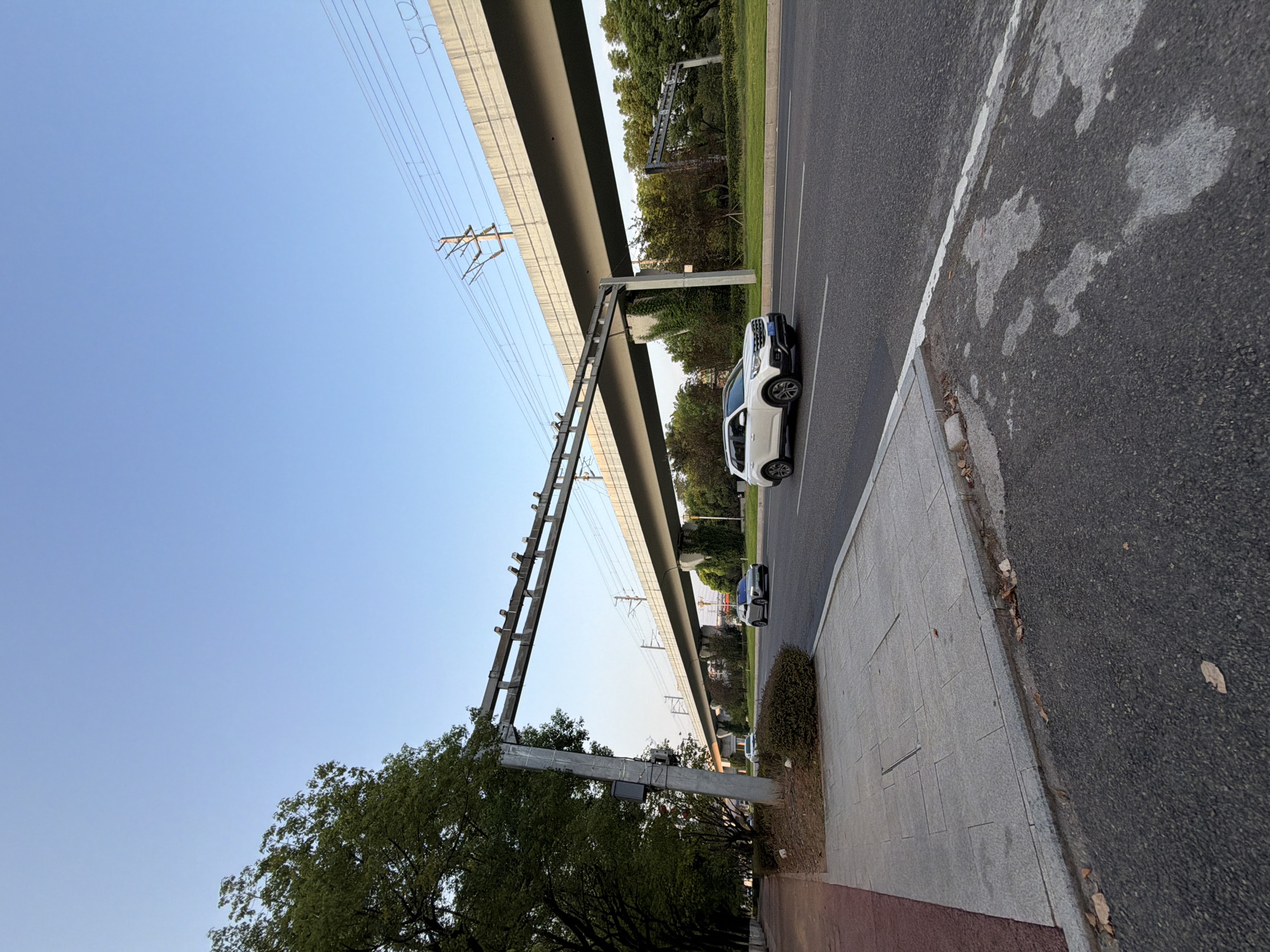The image captures a wide-angle view of a road in Jiangjin District, Chongqing, China, under a clear blue sky. The scene depicts an elevated roadway or bridge structure with visible overhead power lines, possibly for a light rail or train system, spanning across the road. On the right side of the road, a white SUV is in motion, driving on the asphalt. To the left of the road, a sidewalk is visible, bordered by trees that form a dense canopy. The overall environment suggests an urban or suburban setting with infrastructure for transportation.