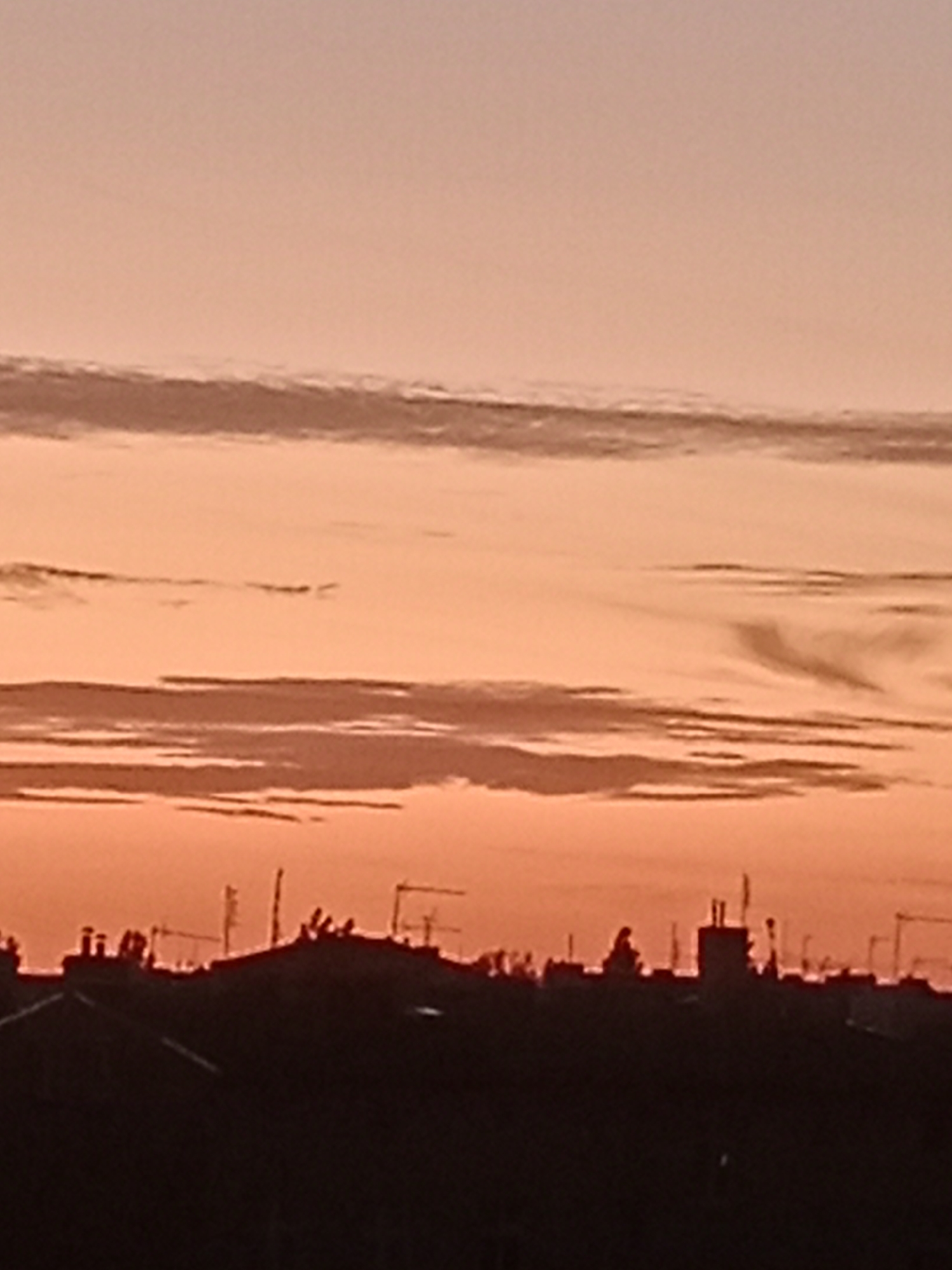 A silhouette of a cityscape is visible against a twilight sky. The sky is a gradient of soft peach and pale orange, with wispy clouds layered horizontally across the frame. The lower portion of the image is dominated by the dark silhouette of buildings, rooftops, and various antennas and structures. The skyline suggests an urban environment. While the exact location in Bydgoszcz, Poland, cannot be determined from the visual information alone, the presence of dense urban architecture with antennas is characteristic of many cityscapes. No individuals are clearly discernible within the silhouette, only the general outlines of structures.