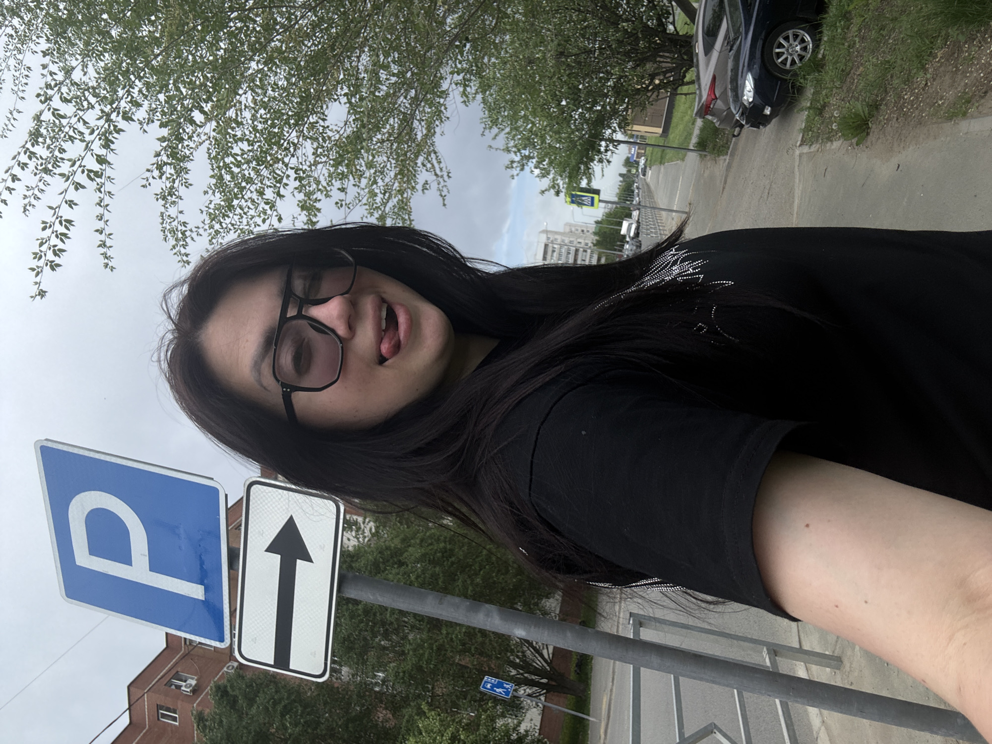 A selfie taken in a city environment. A person with dark hair and glasses is visible. The person is wearing a black shirt and has their tongue slightly sticking out. The background includes a blue parking sign with a white "P" and a white arrow sign pointing straight ahead. Beyond the signs, there is a street with parked cars, trees, and buildings. The sky is overcast. The location appears to be a sidewalk next to a street in an urban area.