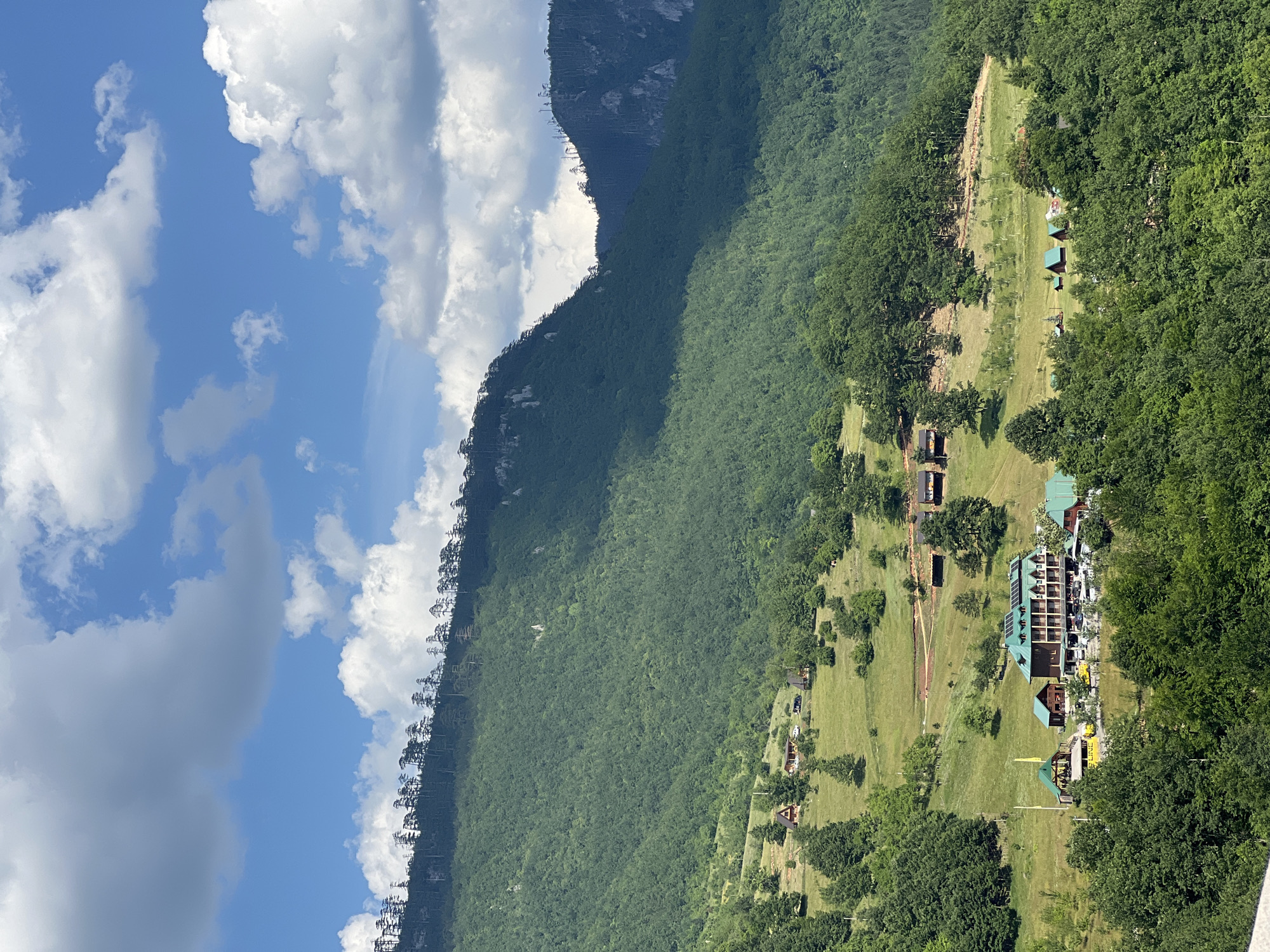This is an aerial, eye-level, long shot of a mountain village in Đurđevića Tara, Montenegro. The scene is dominated by lush green mountains, with a clear blue sky dotted with fluffy white clouds. In the foreground, dense green trees form a natural frame. Below the treeline, the landscape opens up to rolling hills covered in grass and scattered trees.
Several wooden houses with green roofs are nestled into the hills. A larger building complex, also with green roofs and a multi-story structure, sits in the mid-ground, surrounded by parked vehicles. Further up the hill, more scattered houses are visible. The distant mountain slopes are densely forested, with a line of tall, thin trees topping the highest ridge, silhouetted against the sky. The overall impression is one of a peaceful, remote mountain settlement bathed in sunlight.