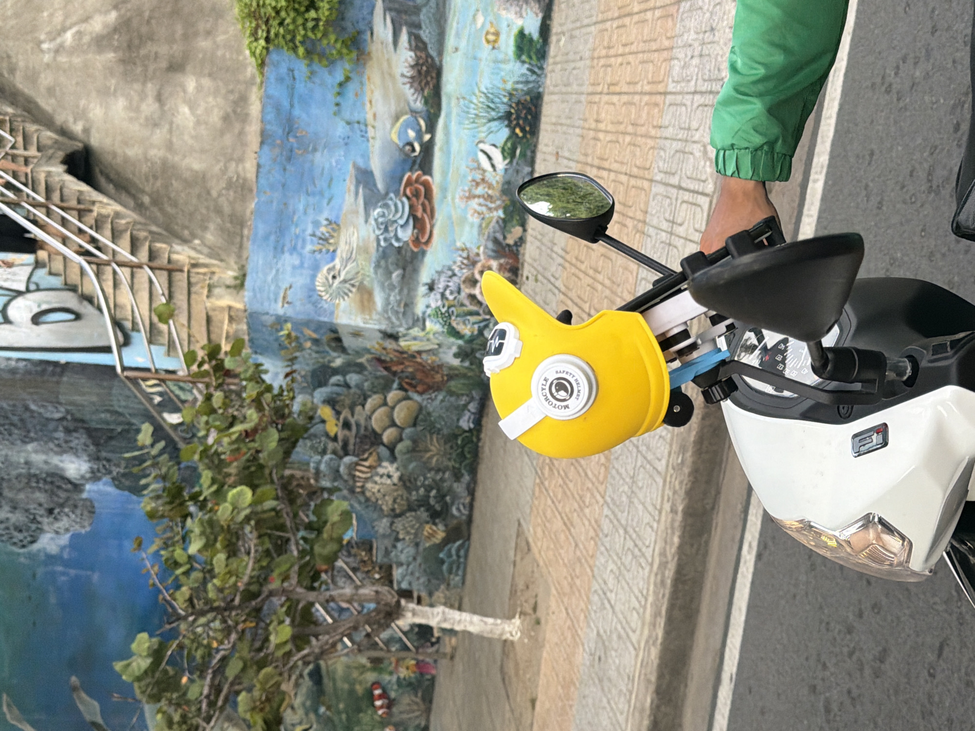 In Nha Trang, Vietnam, a close-up view captures a person's right arm and hand wearing a green jacket, resting on the handlebar of a white scooter. Attached to the handlebar is a bright yellow child's helmet, decorated with a white decal featuring a motorcycle and text. The scooter's mirror reflects lush green trees and a blue sky. In the background, a vibrant mural depicts an underwater scene with coral and fish, adorning a concrete wall. A staircase with a metal railing ascends the wall, suggesting an elevated walkway or building. The ground is paved with patterned tiles and asphalt.