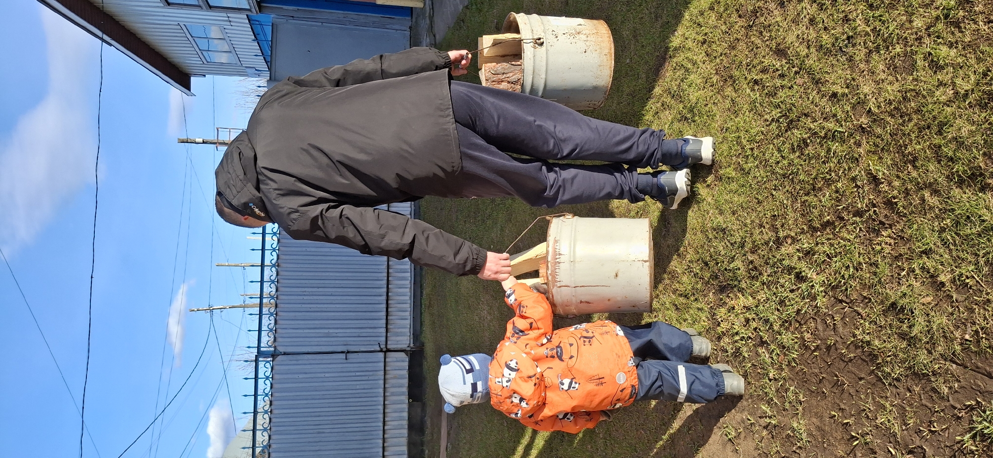In Chaikovsky, Russia, a man in a black jacket and dark pants assists a young child in an orange jacket and blue pants, who is carrying a gray metal bucket filled with wooden planks, using rope handles. Another similar bucket filled with wood is nearby. The scene takes place on a patch of grass and dirt, adjacent to a building with a metal fence and a blue sky with light clouds visible in the background.
