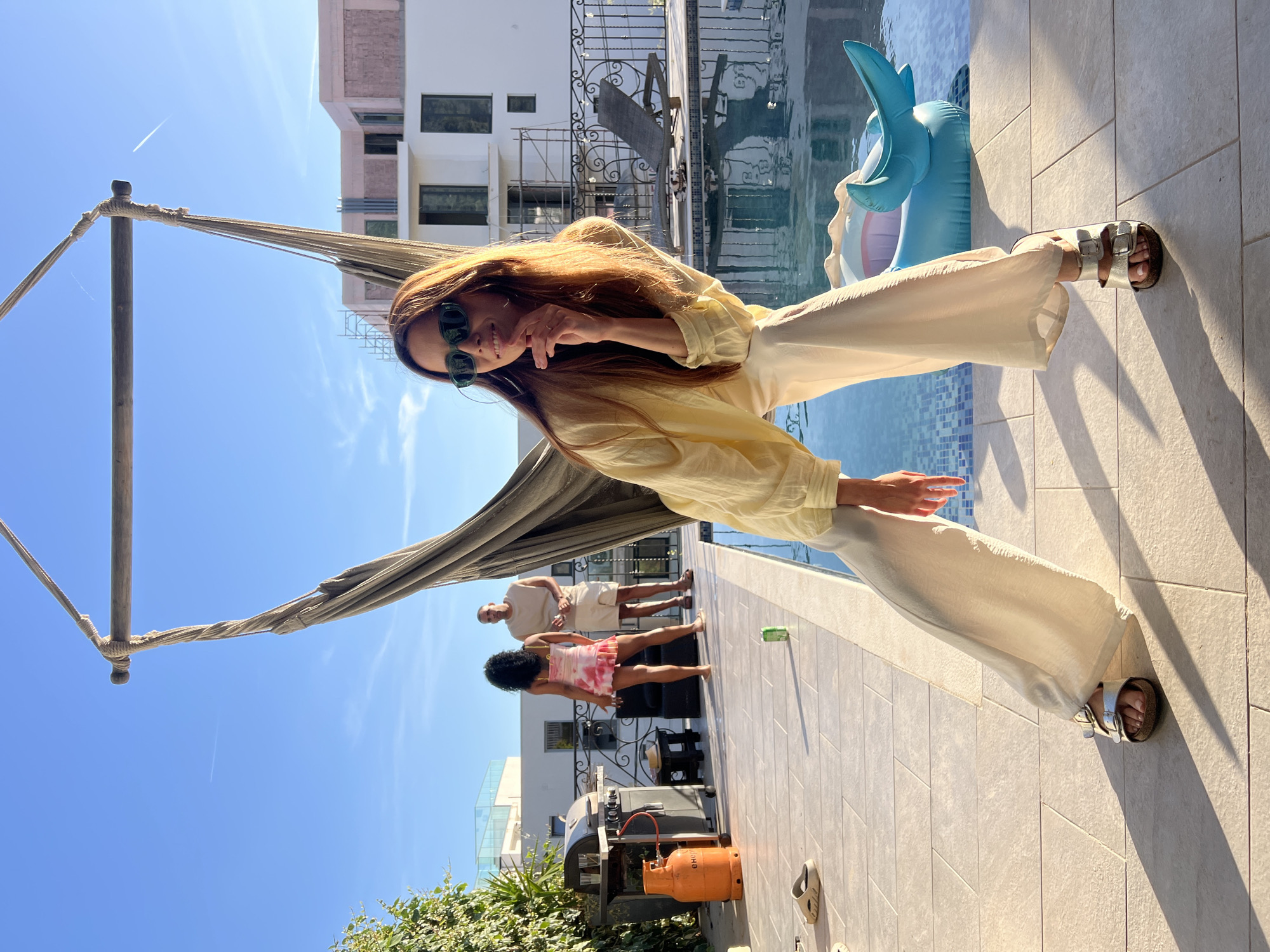 A full shot captures a light-skinned woman with long reddish-brown hair and sunglasses, standing on a tiled patio next to a pool with an inflatable dolphin. She wears a light yellow button-down shirt and light-colored pants, posing with one hand raised and two fingers extended. Behind her, two figures stand on the patio: a light-skinned man in a tan shirt and shorts, and a Black woman in a pink and white dress. A hammock structure is visible to the left, along with a grill, an orange propane tank, and greenery. A multi-story building is in the background, partially obscured by construction scaffolding. The scene is located in Dobra Voda, Montenegro, evidenced by buildings and terrain.