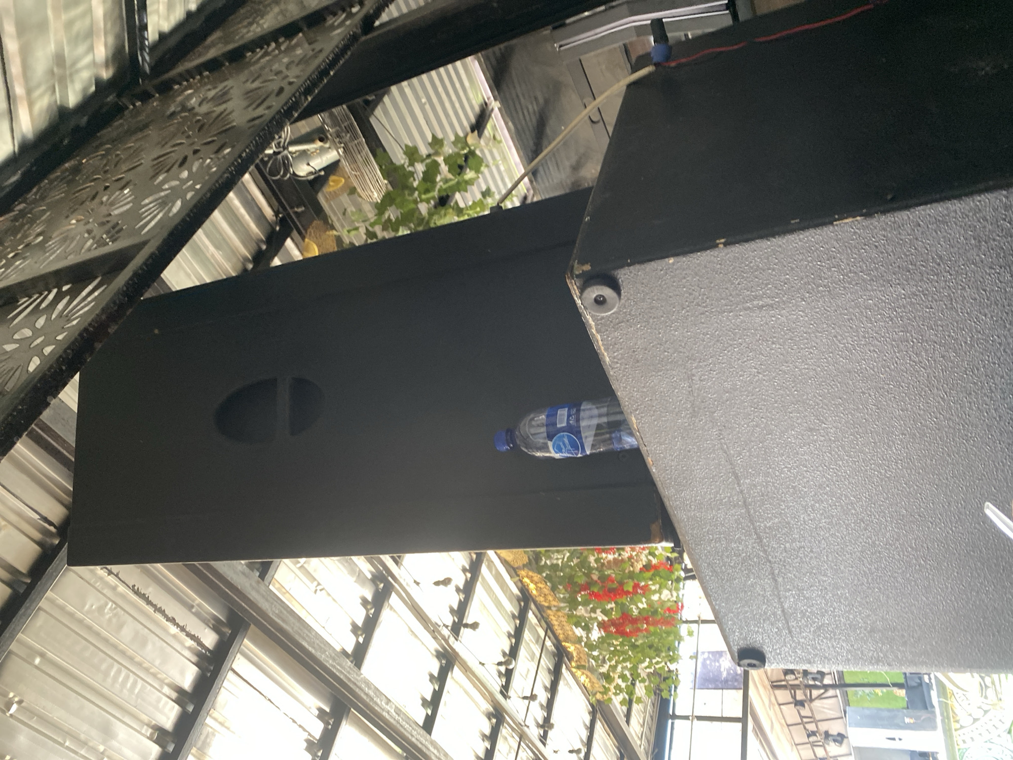 The image presents an upward-angled view within a covered outdoor or semi-outdoor event venue in Unknown, Unknown. Two large, dark-colored professional audio speakers dominate the foreground. The lower speaker is a wide, rectangular cabinet with a textured, speckled dark grey finish, featuring a circular black rubber foot on its top-left corner. Stacked vertically upon this lower cabinet is a taller, narrower matte black speaker, which has a prominent circular, recessed port or horn opening located in its upper section, slightly off-center to the right. A clear plastic water bottle with a blue screw cap and a blue and white "AQUA" brand label is standing on the top surface of the lower speaker, positioned to the right of the taller speaker. A red cable and a blue connector are visible on the right side of the taller speaker, indicating electrical or audio connections, and a light-colored cable snakes from behind the speakers towards the right.
Above and around the speakers, a bright translucent corrugated roof structure is visible, supported by dark metal beams, allowing significant sunlight to filter into the space. On the upper right, a decorative dark metal panel with intricate, laser-cut floral or leaf patterns extends diagonally across the frame. To the left, artificial green foliage and clusters of bright red decorative flowers are suspended from the roof structure, contributing to the ambiance. In the distant background on the left, additional event equipment, possibly scaffolding or another large speaker unit, is faintly visible. The scene is devoid of people.