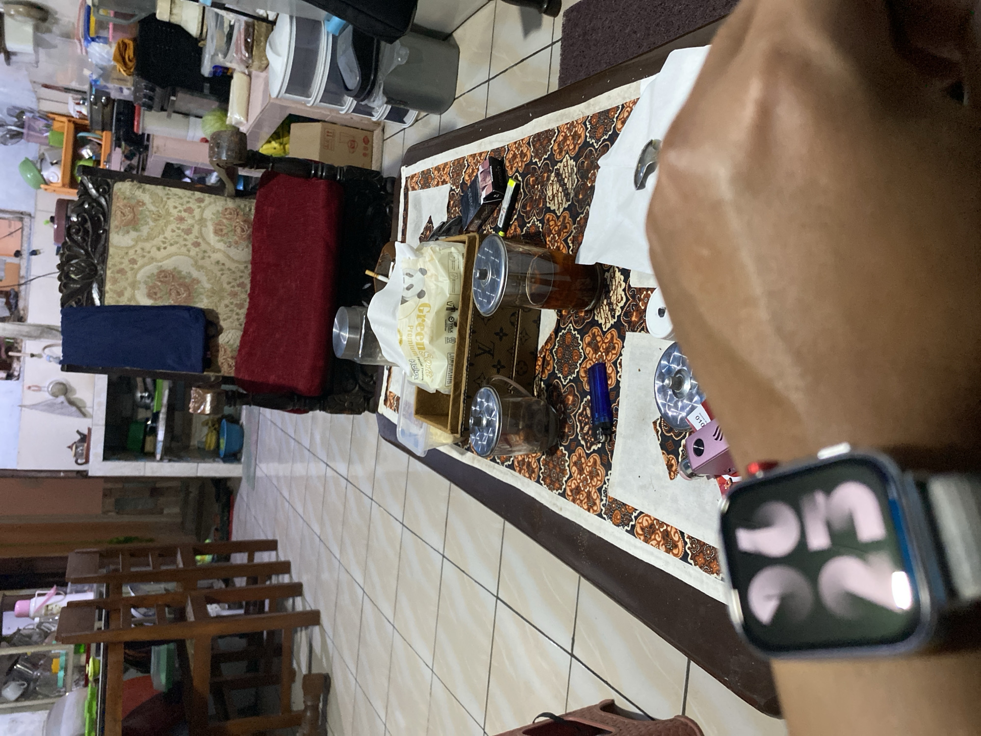 The image shows a close-up of a person's wrist wearing a smartwatch, with a table laid out in the background. The watch displays the time "06:23". The table has a patterned cloth runner with motifs of brown, orange, and white. On the table, there are several objects including a pack of tissues, two glass containers with lids, one filled with dark liquid and another with a dark solid substance, a blue lighter, and a pack of cigarettes. In the background, there is a wooden chair with an upholstered seat and backrest, covered with a red cloth. Further in the background, a shelf unit is visible, holding various kitchen items and containers. The floor is tiled with square, light-colored tiles. The overall setting appears to be a casual indoor environment, possibly a dining area or living room, in a residence in Wonosari, Indonesia.