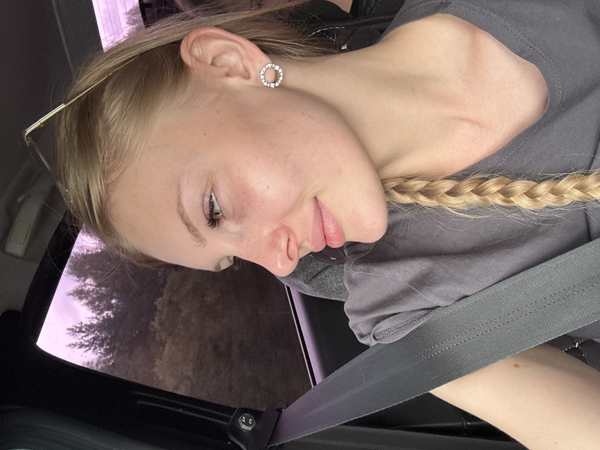 A young blonde woman with fair skin, freckles, and green eyes is seen in profile, looking out of a car window. She is wearing a grey t-shirt and a seatbelt. Her hair is in a braid, and she has a circular earring with small stones on her right ear. In the background, through the car window, a blurry image of trees and a pinkish sky is visible. The image appears to have been taken during daytime, possibly in the city of Shumikha, Russia.
