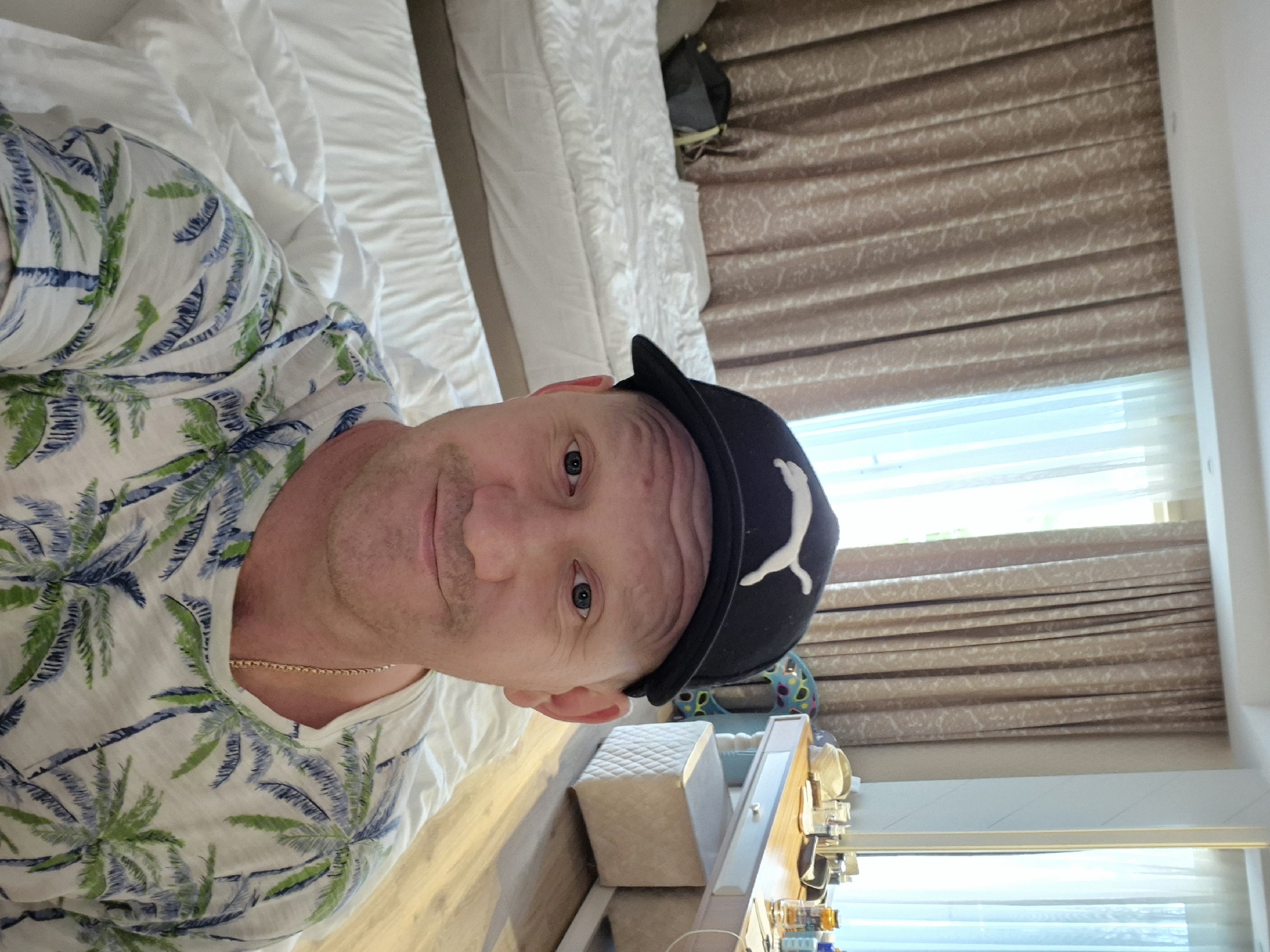 A man wearing a palm-tree-print shirt and a black Puma baseball cap is lying on a bed with white bedding. He has fair skin, blue eyes, and a gold chain. In the background are a bedside table, various items on the table, and a window with brown fabric blinds. The location is likely a hotel room in Alanya, Turkey, given the context.