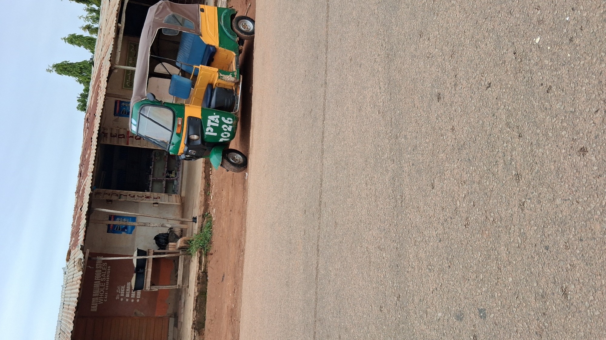 In Jos, Nigeria, a green and yellow auto-rickshaw, with "PTA 1026" painted on its side, is parked on the shoulder of a paved road. The auto-rickshaw has a brown canopy and blue seating. Behind it, a corrugated metal-roofed structure serves as a storefront. Signs on the storefront indicate it is a "HAJIYA HALIMA FOOD STORE WHOLE SALES" and lists items for sale such as rice, beans, and groundnuts, along with a phone number for inquiries. The ground is a mix of reddish-brown dirt and sparse green vegetation. The sky above is overcast with clouds.