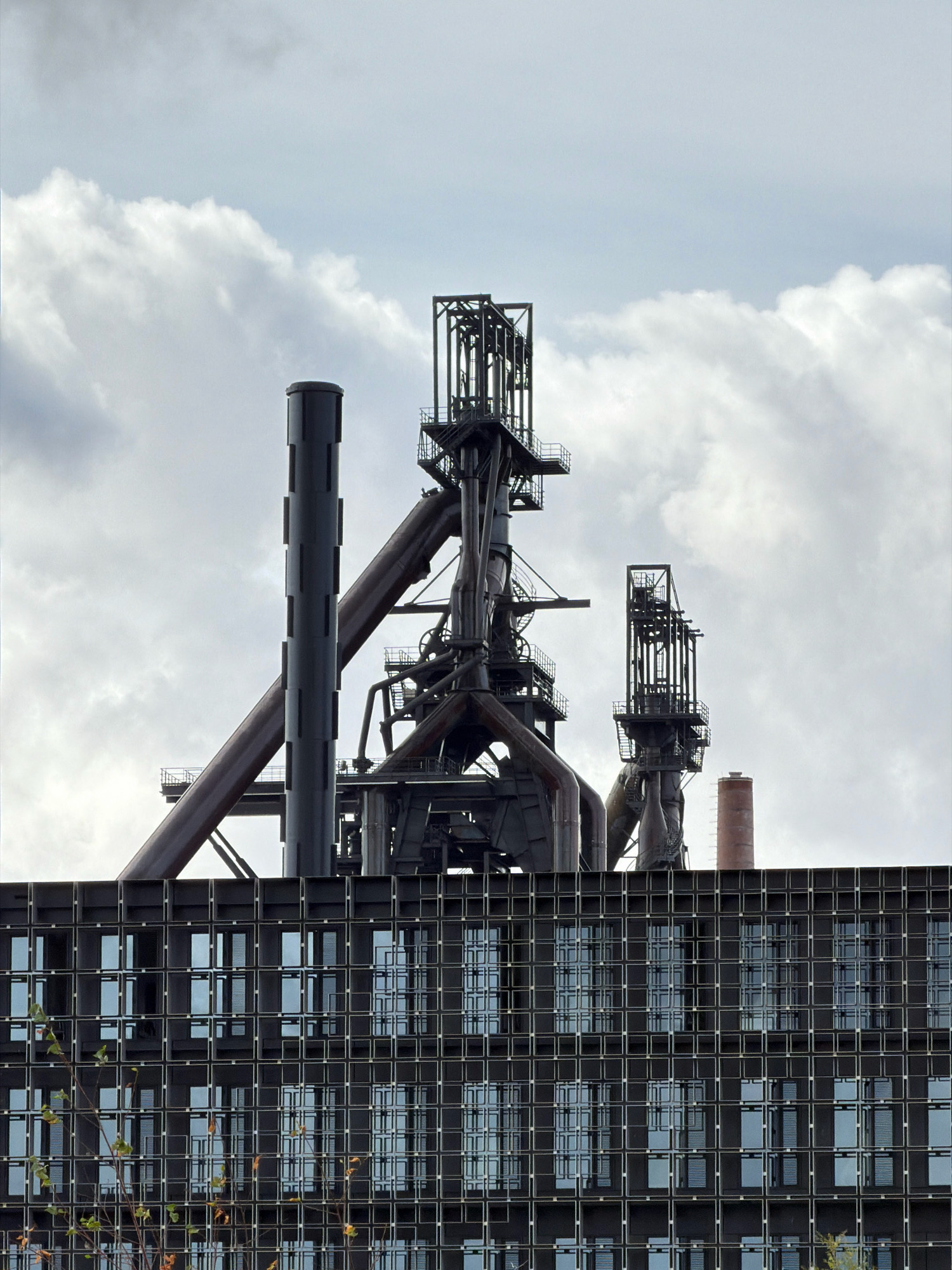 The image depicts the industrial landscape of Esch-Belval in Esch-sur-Alzette, Luxembourg. Dominating the upper half of the frame are towering, dark metal structures of a former blast furnace complex. These industrial remnants feature intricate scaffolding, large pipes, and chimneys against a backdrop of a cloudy sky. In the foreground, a modern, dark building with a grid-like facade of numerous windows stretches across the lower portion of the image, contrasting sharply with the aged industrial machinery above. Some bare branches of a tree are visible in the lower left corner, hinting at the changing seasons.