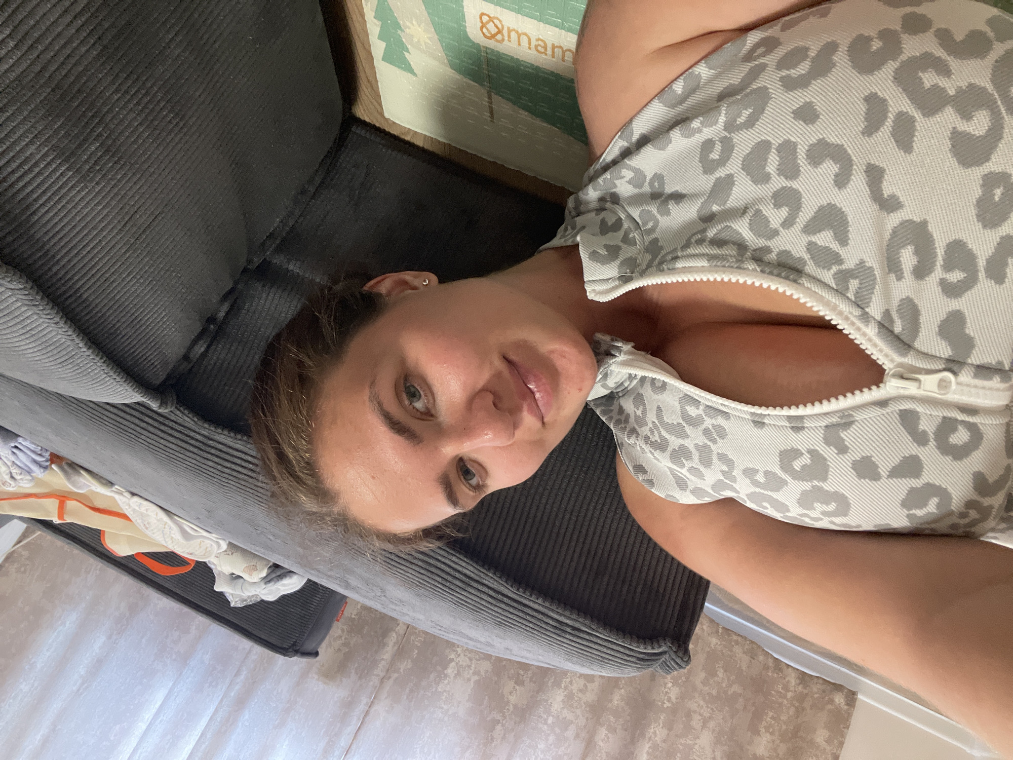 A woman is lying on a gray, corduroy sofa. She is wearing a light gray and white leopard-print top with a zipper. A "mam" branded mat is partially visible next to the sofa. The location of the scene is not discernible and there is no evidence to suggest that it is in Misaylovo, Russia.