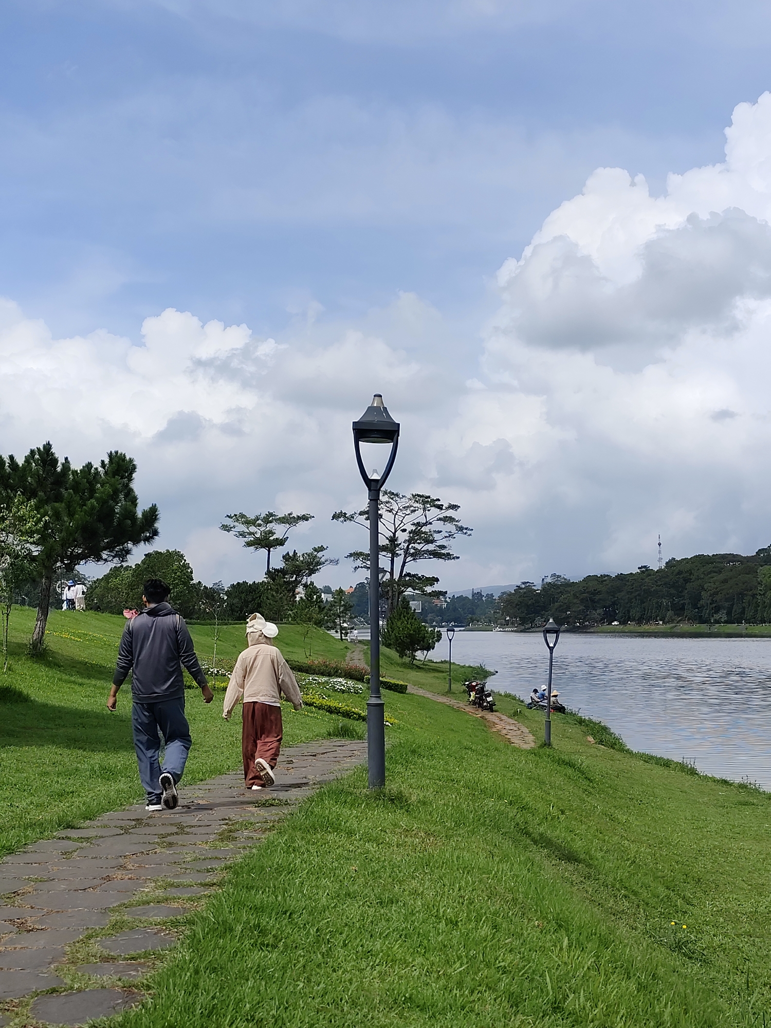 The image presents a serene outdoor scene, characteristic of a park area by a lake in Da Lat, Vietnam. A tranquil body of water, likely a lake, occupies the right side of the frame, its surface gently rippled under a bright sky. A winding paved pathway, constructed from irregular stone slabs, guides the eye through lush green grass that slopes down towards the water's edge. Beyond the lake, densely forested hills rise, with a distant slender tower just visible above the tree line. Scattered pine trees and other green foliage dot the landscape on both sides of the water.
In the foreground, a man and a woman walk away from the viewer along the stone path. The man wears a dark grey hoodie and blue jeans, while the woman is dressed in a light-colored hooded top and reddish-brown wide-leg pants, with a head covering. Their relaxed posture suggests a leisurely stroll. Several dark lampposts with distinctive tops are spaced along the pathway, leading into the distance. Further along the lake's bank, two individuals are seated on the grass, appearing to relax or fish, with two motorcycles parked nearby. More blurry figures can be seen on a grassy hill to the left, indicating other people enjoying the park.
The sky is bright blue with scattered white and light grey clouds, suggesting pleasant weather conditions during the daytime, likely late morning or early afternoon. The scene evokes a sense of peace and leisure, highlighting the natural beauty and recreational opportunities found in Da Lat. There is no visible text in the image.