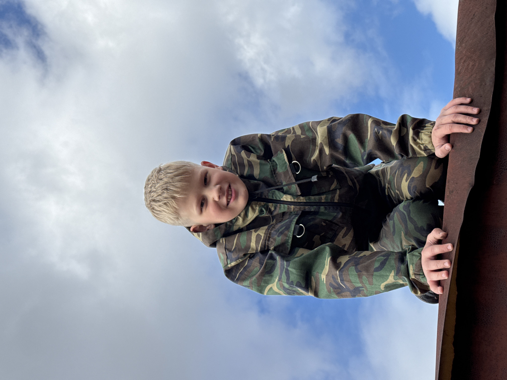 A young child with blond hair, wearing a camouflage-patterned jacket and pants, is shown from a low angle against a background of a cloudy sky. The child is holding onto the edge of a rusted metal structure. The location may be Gornyi (Dvurechenskoe S/P), Russia.