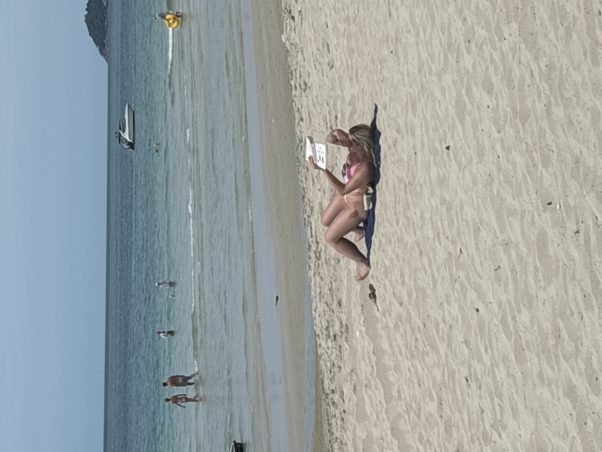 A person is lying on a sandy beach, reading a book. The person is wearing a pink bikini and is lying on a dark towel. The beach is a light tan color with small ripples in the sand. In the background, there is a body of water with several people wading and swimming. A white boat is visible on the water. The sky is a light blue with some clouds. The overall scene depicts a relaxing day at the beach.