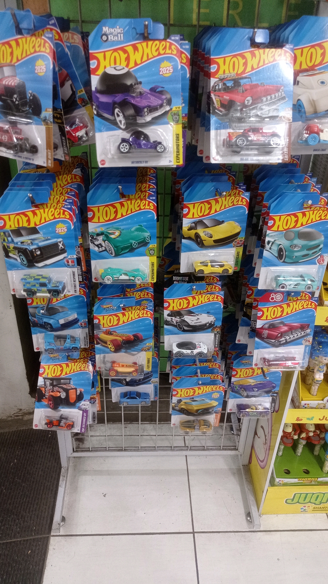 The image shows a retail display of Hot Wheels toy cars. The display features multiple rows of blister-packed toy cars. The packaging is primarily blue with the Hot Wheels logo and images of the cars. Some of the cars shown are purple, red, green, yellow, and blue. The display rack is made of white metal. In the background, there is a green wall and a portion of another display with toy figures. The image appears to be taken at a retail store. The presence of a Hot Wheels display suggests a toy store, department store, or supermarket with a toy section. The specific location of Mulyoagung, Indonesia, cannot be determined from the image alone, as there are no identifying landmarks or signage.