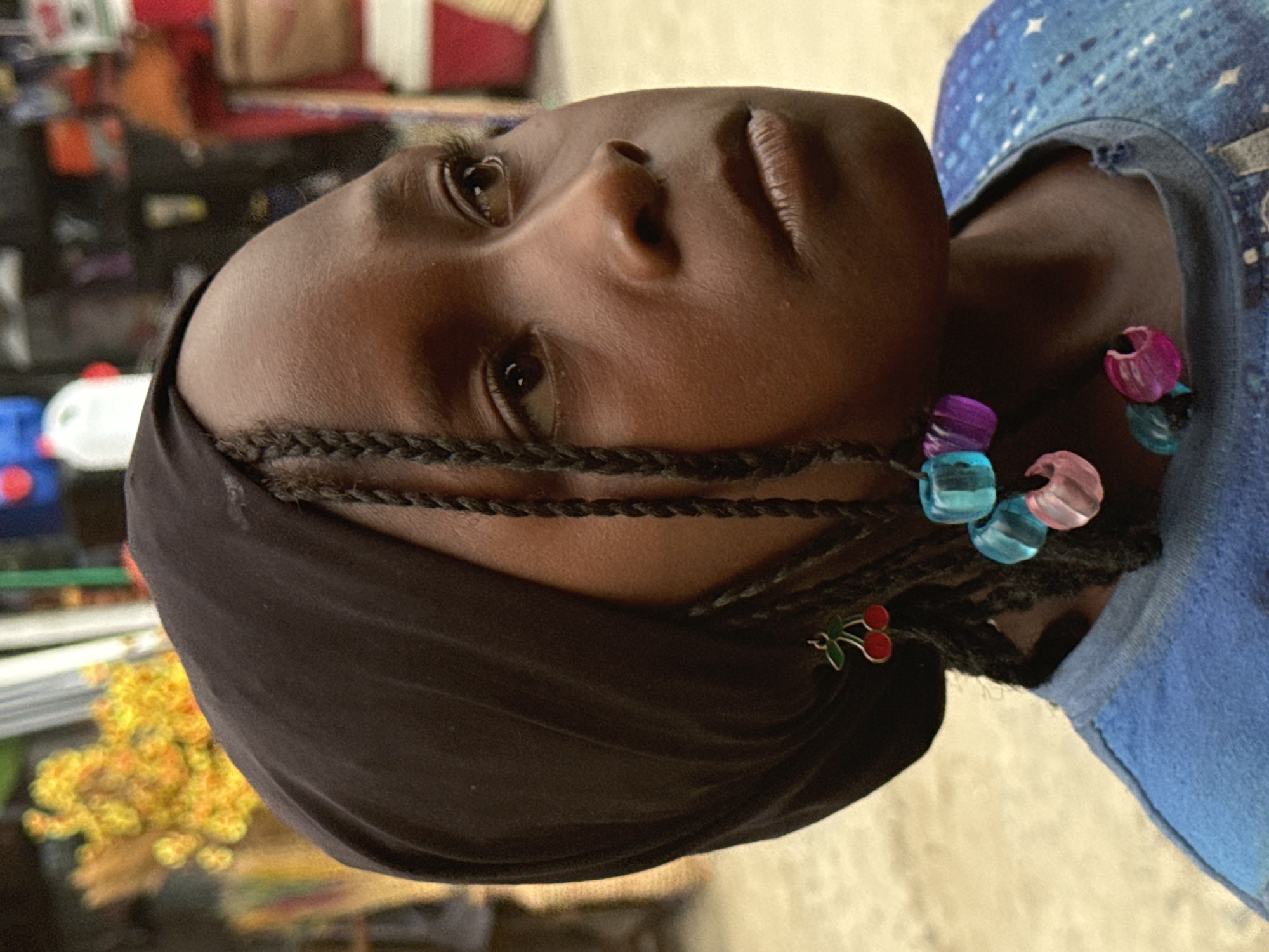 A close-up of a young, dark-skinned individual with braided hair adorned with colorful beads and a cherry charm. The person is wearing a dark head covering. The individual appears to be somewhere in Lagos, Nigeria, based on context clues in the image, but I am unable to pinpoint an exact location without other information.