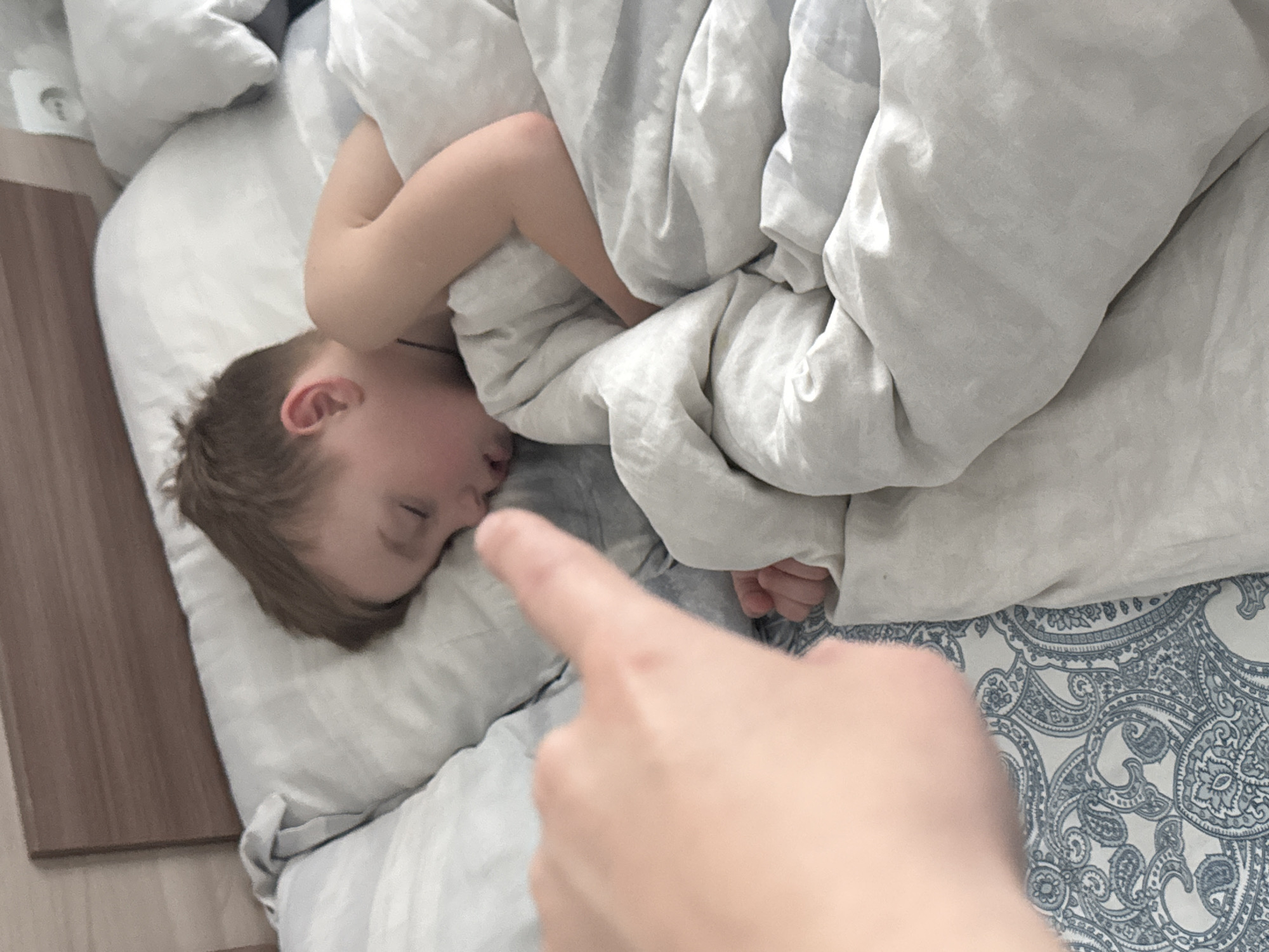 A high-angle close-up shot captures a serene moment with a young child sleeping soundly in a bed. The child, likely a boy with light brown hair, lies on his side on a light grey pillow, eyes closed in slumber, partially covered by a plain light grey duvet. One arm is tucked close to his head beneath the covers.
In the foreground, a fair-skinned hand, presumably an adult's, extends towards the child, with an index finger gently pointing at or near the child's nose. This gesture suggests a moment of tender interaction, observation, or perhaps a playful attempt to rouse the sleeping child.
The bedding consists of light grey pillows and a matching duvet. Below the duvet on the lower right side, a white bedsheet with an intricate blue and grey paisley pattern is visible. To the left of the bed, there is a dark wooden surface, possibly part of the bed frame or furniture, next to a lighter wooden floor. The lighting in the room appears soft and diffused, indicating a calm indoor setting, likely during the daytime. The scene portrays a quiet, intimate moment within a home in Chaikovsky, Russia. No text is visible in the image.