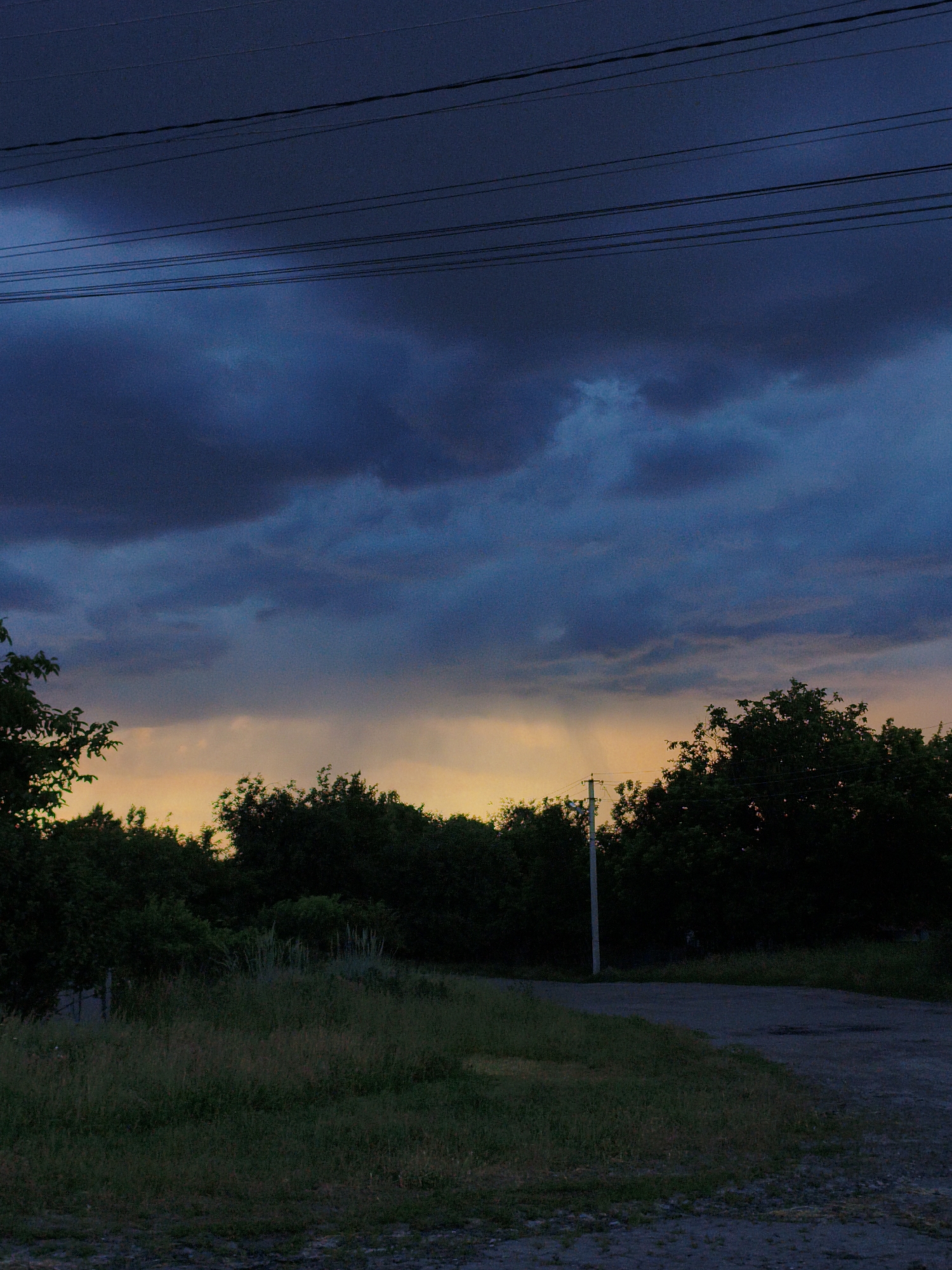 The image depicts a rural scene at dusk in Ustyvytsia, Ukraine. In the foreground, a gravel path curves from the right towards the left, bordered by overgrown grass and vegetation. A dirt road is visible on the right side of the frame. A single utility pole stands in the middle ground, silhouetted against a backdrop of dense trees that form a dark line across the horizon. Above the trees, the sky is a dramatic mix of deep blues and grays, with dark, heavy clouds suggesting an impending storm. A band of muted orange and yellow light is visible near the horizon, indicating the last remnants of sunset breaking through the clouds. Several power lines are stretched horizontally across the upper portion of the image, adding to the utilitarian feel of the rural landscape. No people or animals are present.