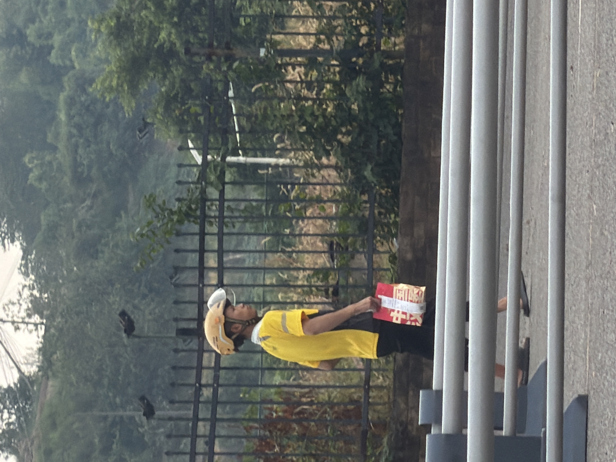 A person wearing a yellow shirt, black pants, and a yellow helmet with a clear visor walks along a paved road. They are carrying a red and white package in their right hand. The person is walking on the right side of the frame, facing towards the left. Behind them is a black metal fence with vertical bars, beyond which is a grassy hill with trees and some structures that appear to be lighting poles. In the foreground, there are several horizontal gray barriers. The scene appears to be in a city, possibly in Jiangjin, China, given the context. The overall lighting suggests it is daytime, with overcast or hazy conditions.