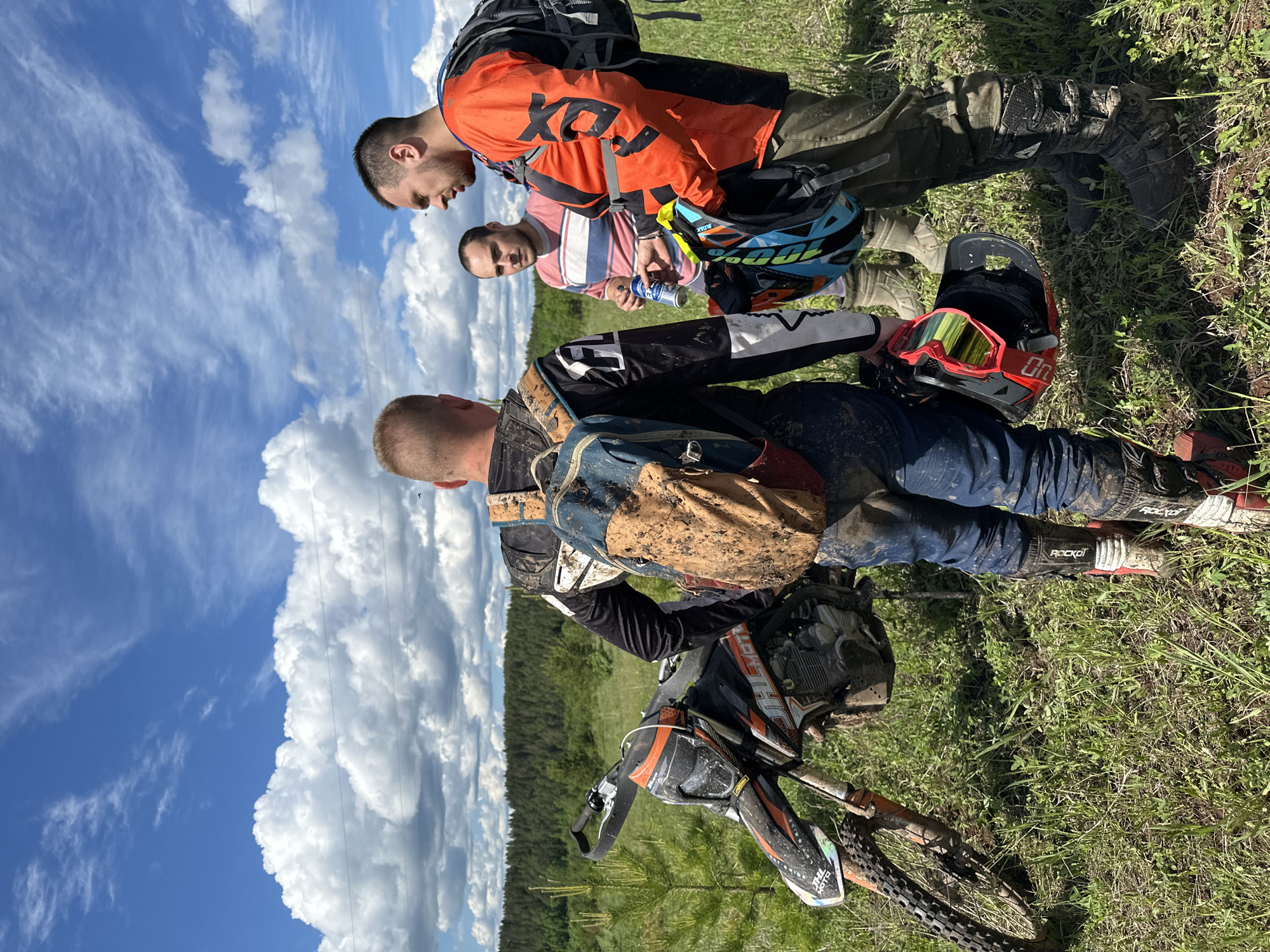 Three men are standing in a grassy field with a dirt bike. The man closest to the camera is lying on the ground next to the dirt bike, which is covered in mud. He has a backpack on and is wearing riding gear, including boots with "Rocko" written on them. The other two men are standing next to him, looking down. One man is wearing an orange and black Fox Racing jersey and khaki pants, and the other is wearing a pink and white striped shirt. A blue and black helmet and a red helmet with goggles are also on the ground. A forest is visible in the background, and a blue sky with white clouds is overhead. The location is Пепеляева, Russia.