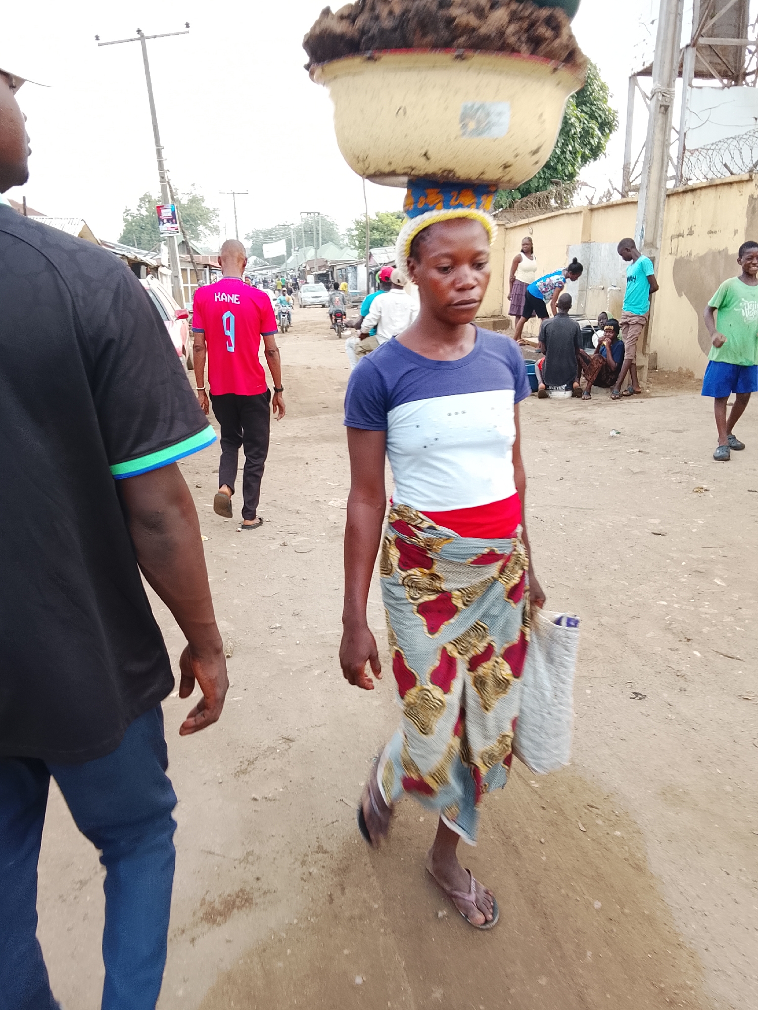 The image shows a woman walking on a dirt road in Abuja, Nigeria. She is carrying a large bowl on her head and holding a bag in her hand. She is wearing a blue and white shirt, a colorful patterned wrap skirt, and sandals. Other people are visible in the background: a man in a pink jersey labeled "KANE 9" walking in the same direction, a group of people gathered near a wall, and another man in a green shirt walking toward the camera. The setting appears to be a lower-income neighborhood, and the road seems unpaved. There are power lines and various buildings visible in the background. The overall scene suggests a bustling yet modest street environment.