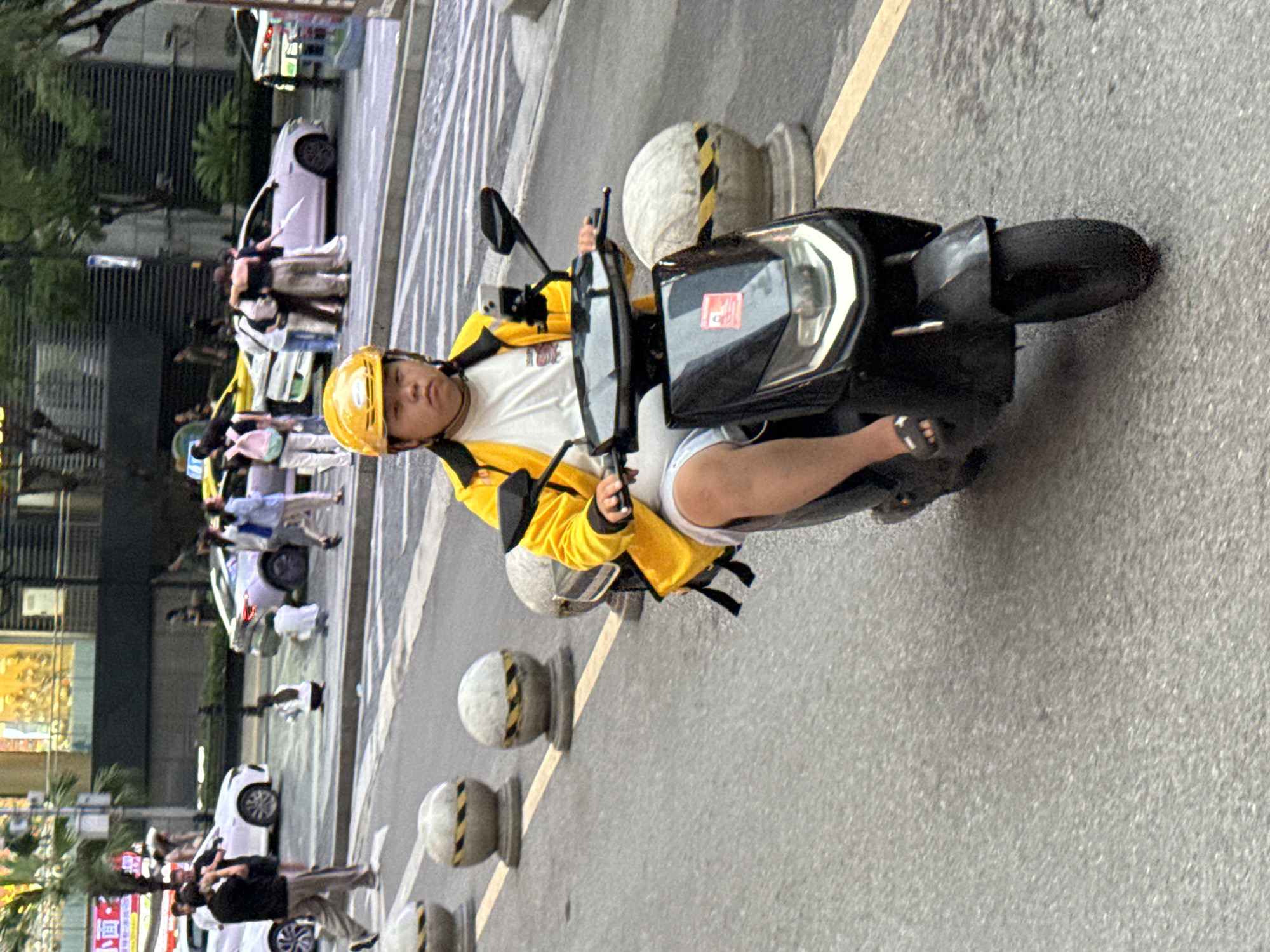 A person wearing a yellow helmet and a yellow jacket rides a black electric scooter on a city street in Yuzhong District, China. The scooter has a black front panel with a red sticker and a phone mount holding a smartphone. The rider is wearing a white t-shirt and light-colored shorts, with dark sandals. In the background, other people are visible, as well as several white cars, one of which appears to be a minivan, and a purple car. The street is paved with asphalt and marked with white and yellow lines. Concrete bollards with yellow and black stripes are positioned along the side of the road. Buildings with windows and signs are visible in the far background. The overall scene depicts a busy urban environment with traffic and pedestrians.