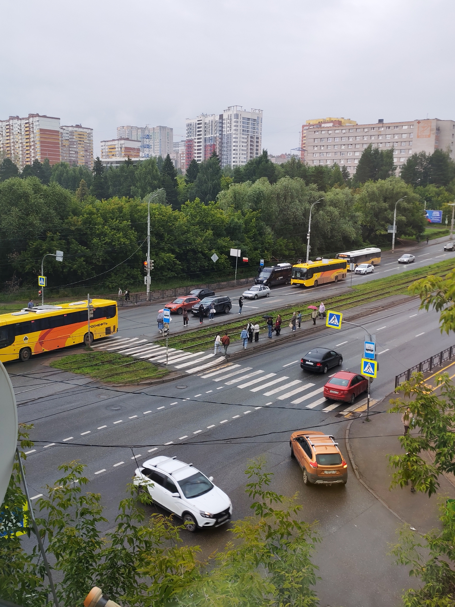 This media file shows a street scene in Izhevsk, Russia, likely on a cloudy, overcast day with some rain. The scene captures a busy intersection with several vehicles, including a yellow bus with "РОСНЕФТЬ" visible on its side, a white SUV, an orange SUV with a roof rack, a red sedan, and a black sedan. Pedestrians are visible near crosswalks, some holding umbrellas, indicating potential rain.
In the background, several multi-story residential buildings and some trees line the street. The buildings appear to be typical of urban architecture in Russia. The street is wet, suggesting recent rainfall, and the sky is a uniform gray. Traffic lights are visible, with one showing a red light. Several blue and yellow pedestrian crossing signs are also present. The overall atmosphere is that of a normal, albeit damp, day in an urban environment. The image appears to be taken from an elevated perspective, possibly from a window or balcony, looking down at the street.