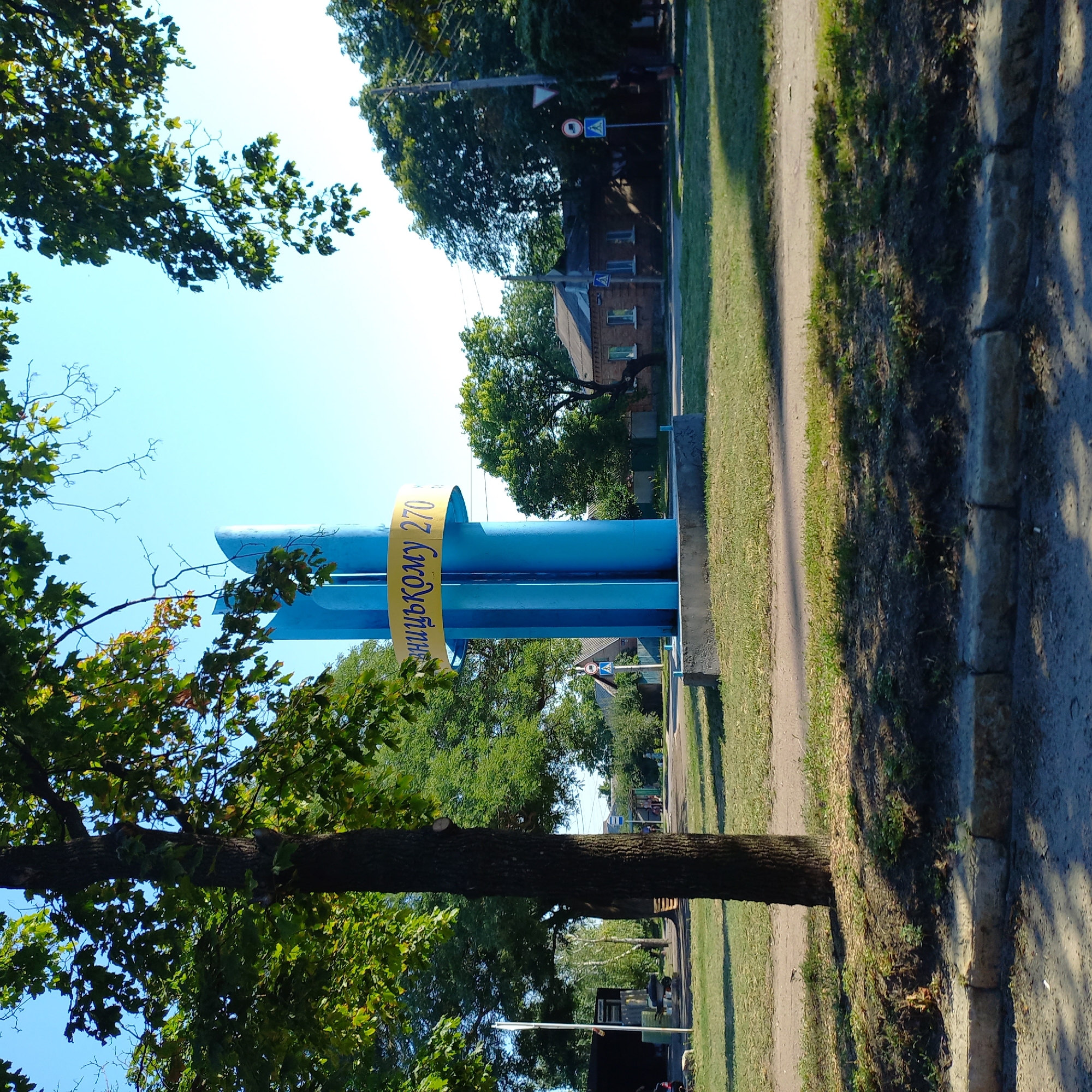 A monument with blue cylindrical structures and a yellow band with Ukrainian text is visible in Kropyvnytskyi, Ukraine. The text on the yellow band appears to be "РІЧНИЦЬКОМУ 270", which translates to "Anniversary 270". The monument is situated in a grassy area, with trees and a clear blue sky in the background. Several houses, a road, a bus stop, and a traffic sign are also part of the scene. A tree trunk is prominent in the foreground. The overall setting suggests an outdoor public space.