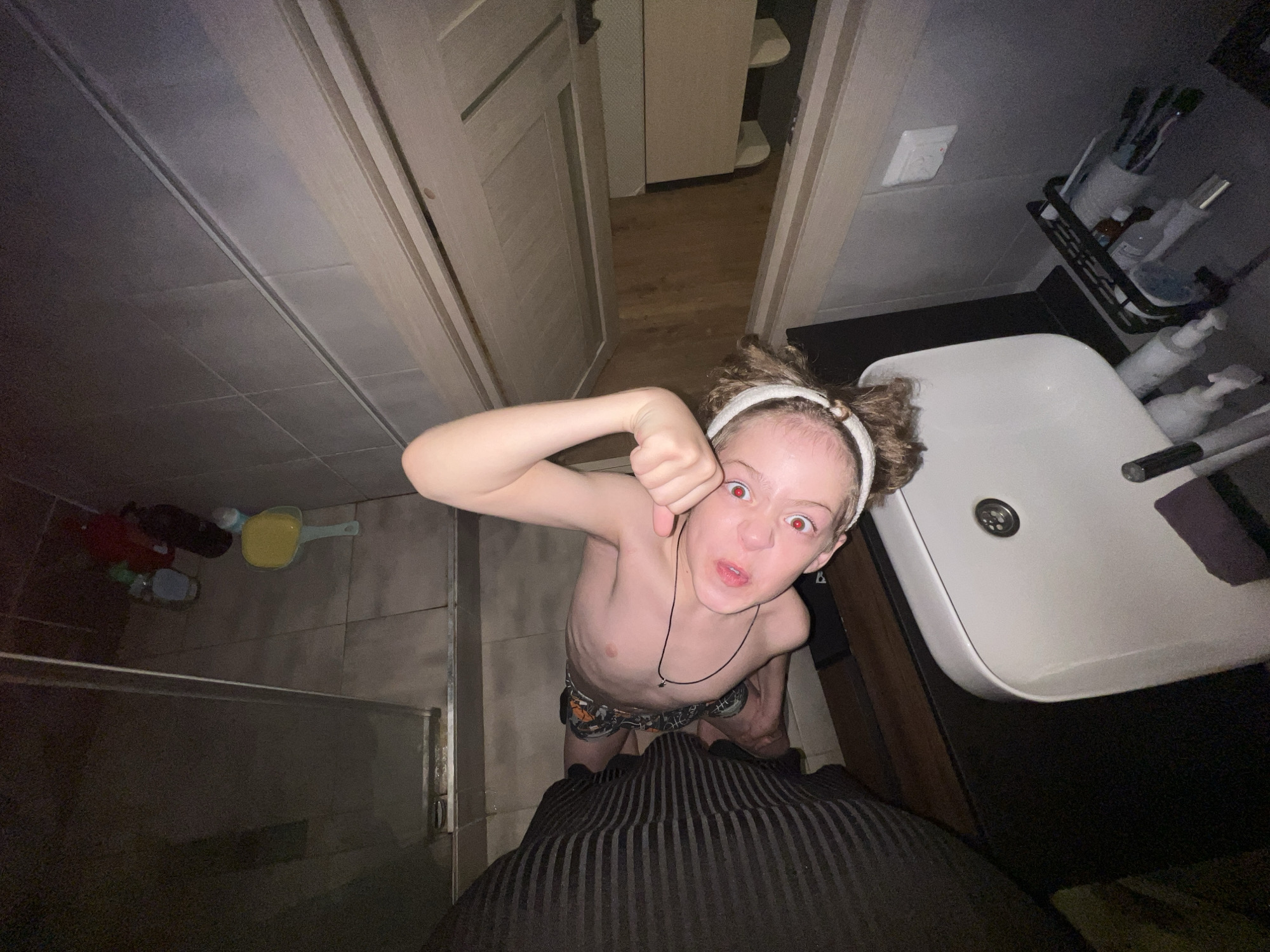 A high-angle shot depicts a child standing in a bathroom. The child has fair skin, reddish eyes, and short, curly hair. They wear a white headband and a necklace. The child is shirtless and wears patterned underwear. Their right arm is raised with a clenched fist. A sink, toiletries, a shower stall, and a door are visible in the background. The bottom part of the image is obstructed by a dark, striped material, likely clothing. The bathroom appears to be located in a modern residential setting. The image was taken in the city of Noviy, Russia.