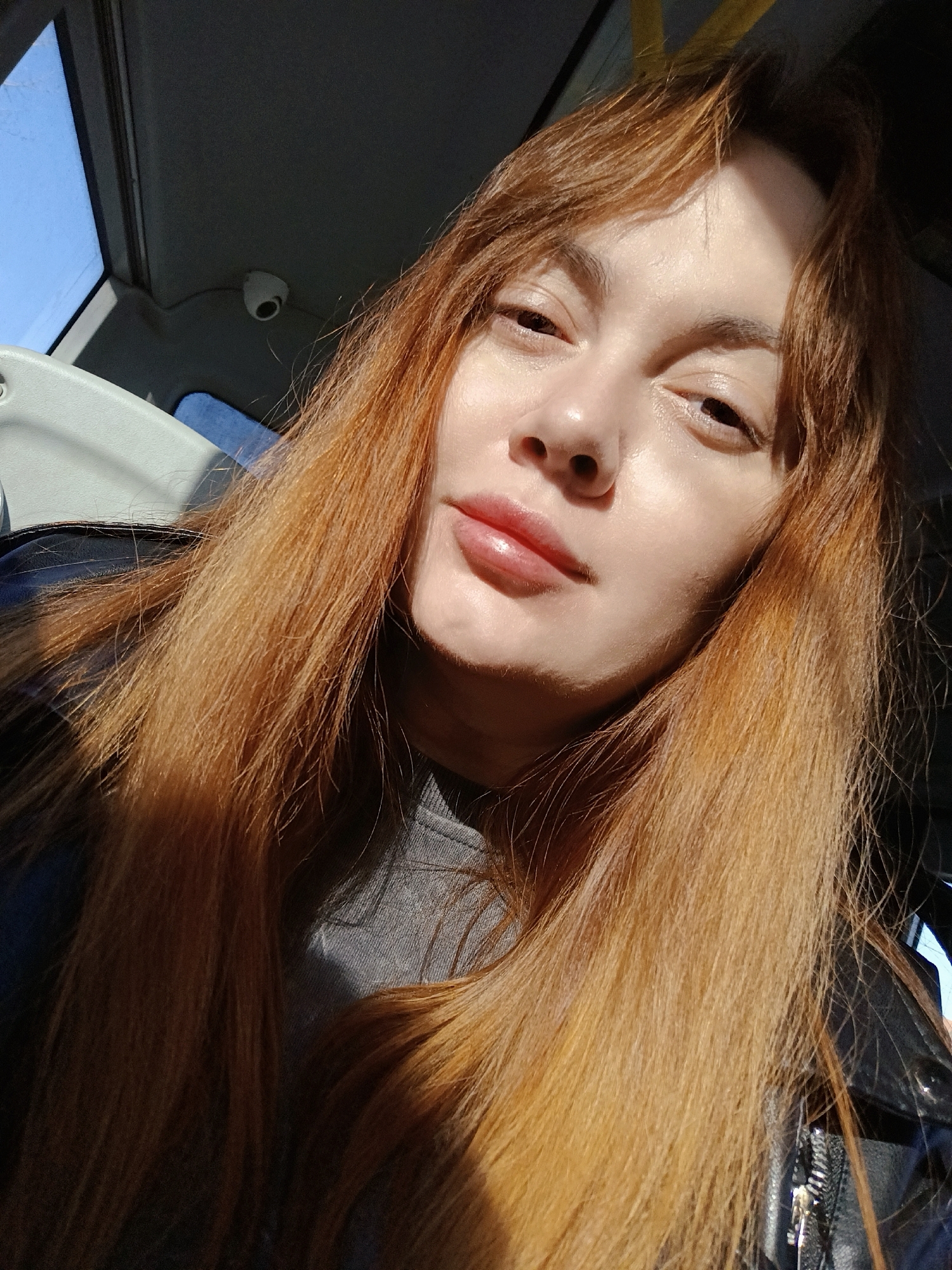 The image is a close-up selfie of a light-skinned person with long, reddish-brown hair. The person is wearing a gray shirt and a dark-colored jacket. The lighting is bright, possibly sunlight, casting shadows on the face and hair.
The location appears to be inside a vehicle, possibly a bus or van, indicated by the presence of a window, ceiling, and what looks like a security camera. The window shows a clear blue sky. Without additional information, definitively identifying the city as Chaikovsky, Russia is impossible.