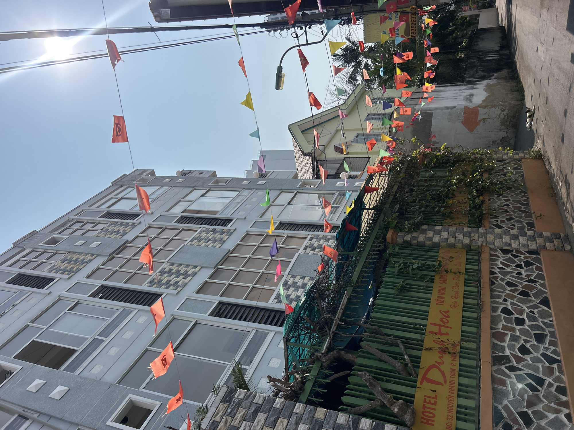 Here is a detailed description of the image:
**Overview:**
The image depicts a street scene in Bắc Nha Trang, Vietnam, showcasing a mix of urban architecture and festive decorations. The photo is taken from a lower angle, looking up towards the sky.
**Buildings and Architecture:**
A modern, multi-story building dominates the left side of the frame. It features a grey facade with numerous windows and decorative stone panels. The building is medium height, approximately 5-6 stories. A smaller, single-story building with a slanted roof is visible behind it on the right, in an off-white color.
**Street and Décor:**
Colorful triangle-shaped flags strung across wires overhead create a celebratory atmosphere. They span the width of the street. These flags include red flags with communist symbols.
**Signage:**
On the right, a sign indicates "HOTEL DƯA HOA" with Vietnamese text providing additional information about the hotel, including the address "ĐC: 32 NGUYỄN KHÁNH TOÀN P. VĨNH HẢ" and contact information. The sign is positioned on a wall with textured stone siding.
**Environment:**
The street appears narrow. Sunlight illuminates the scene, creating a bright and clear sky. The environment suggests a typical Southeast Asian urban setting.
**Elements:**
Green foliage and metal fence-like structures are visible near the hotel sign. The presence of power lines and a street lamp suggest a populated area. The overall image captures a vibrant, urban atmosphere in Bắc Nha Trang.