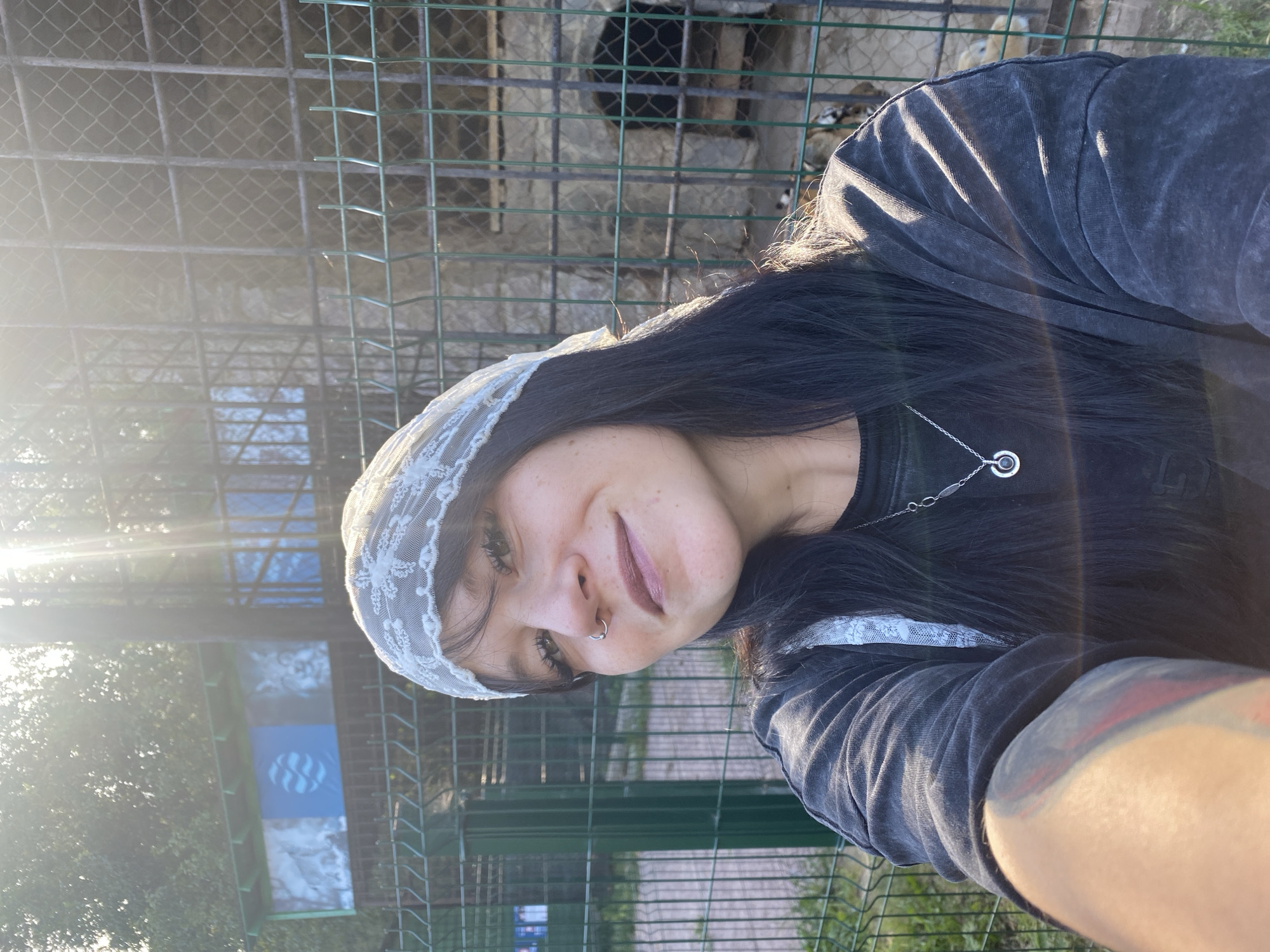 A young woman with dark hair and a white lace headscarf smiles at the camera, holding a smartphone to take a selfie. She is wearing a dark, loose-fitting t-shirt with a silver pendant necklace. A silver hoop nose ring is visible. Her left arm, which is holding the phone, shows a tattoo with red and gray markings.
Behind her, a green chain-link fence encloses an outdoor area. Through the fence, a cage-like structure with metal bars is visible, appearing to be an animal enclosure. In the background, there are trees and a building with colorful panels depicting wildlife. The sunlight creates a lens flare effect across the top portion of the image. The scene appears to be outdoors, possibly at a zoo or wildlife park. The city of Krasnoflotskoe, Russia is not discernable from the visual information provided in the image.
