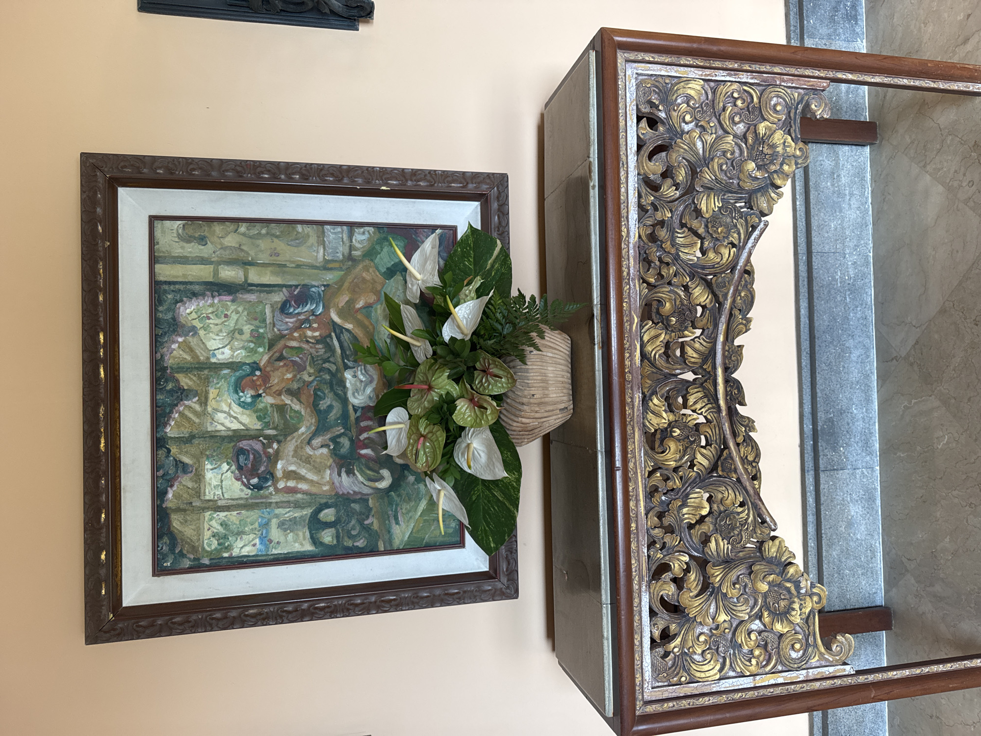 A framed painting of indistinct figures hangs on a pale orange wall, next to a table with ornate gold and dark brown carvings. A vase of white anthurium flowers sits in front of the painting, obscuring part of the artwork. The table has a gray, marble-like top. The location is Kuta Selatan, Indonesia.