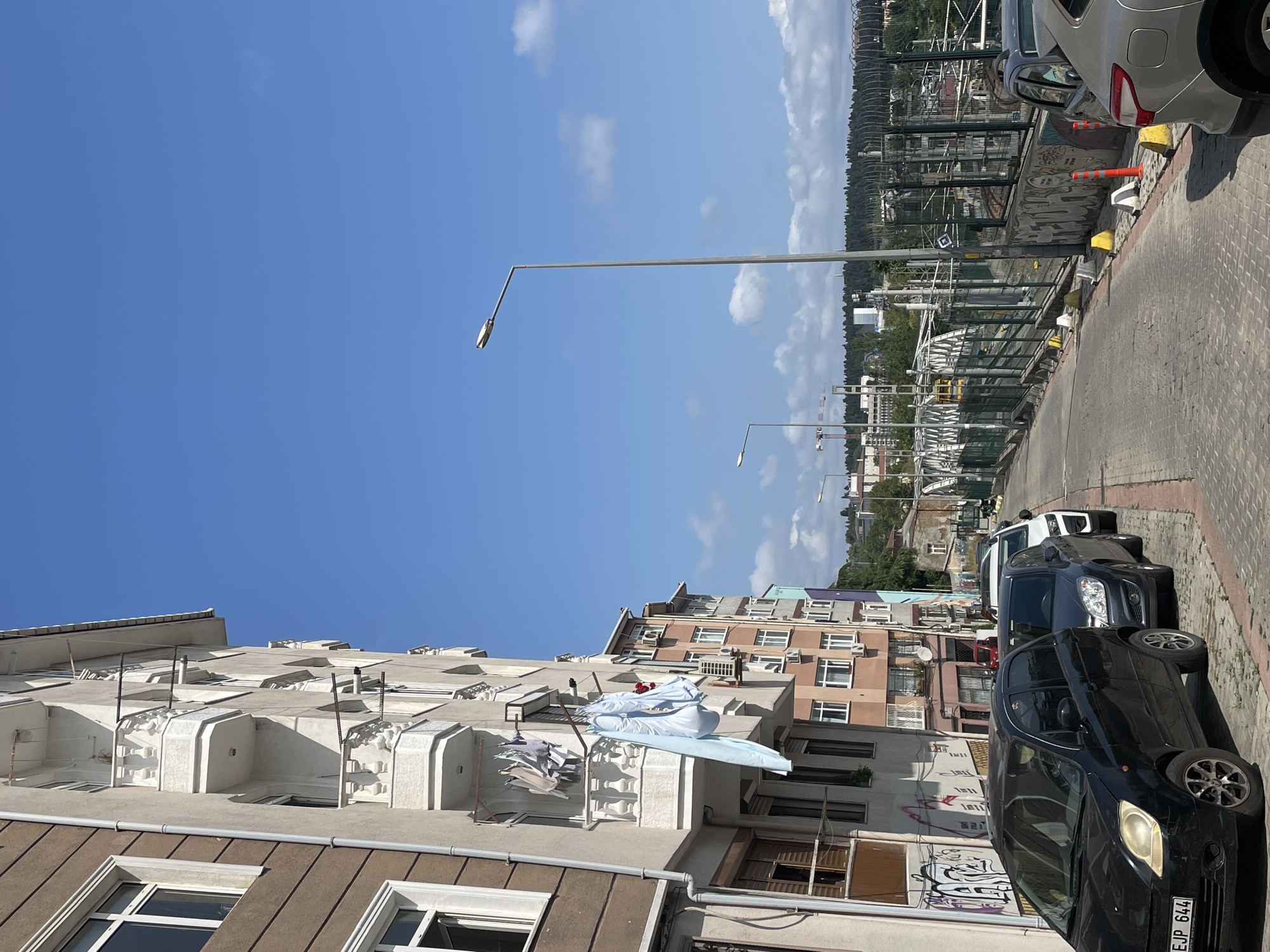 This image depicts a street scene in Kadıköy, Turkey, under a clear blue sky with scattered clouds. On the left side of the street, a multi-story residential building features balconies with white railings and laundry hanging to dry. The building's facade is a mix of brown and light-colored stucco. Adjacent to this building, another residential structure with pink and light-colored walls, air conditioning units, and numerous windows is visible.
The street itself is paved with cobblestones and lined with parked cars, including a black hatchback in the foreground with a visible license plate reading "EJP 644". Behind it, a dark gray hatchback and a white car are parked. The right side of the street is bordered by a metal fence with graffiti on the lower wall. Beyond the fence, a bridge structure with white archways and yellow accents is prominent, suggesting a roadway or railway crossing. Construction cranes are visible in the distance. A tall streetlamp stands near the center of the frame, illuminating the scene. The background consists of a treeline and more buildings, indicating a densely populated urban area. The overall impression is of a sunny day in a residential neighborhood with urban infrastructure.