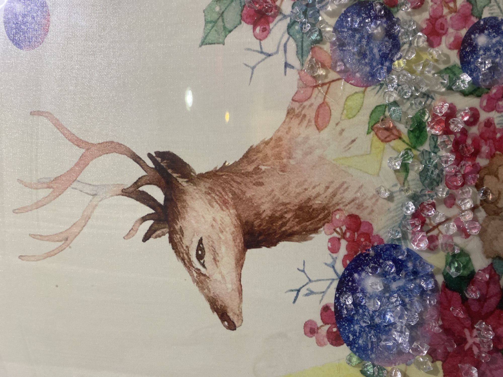 The image depicts a close-up view of a painting featuring a deer. The deer is rendered in a watercolor style, with shades of brown and pink dominating its fur and antlers. The background of the painting includes stylized floral elements in red, green, and pink hues, with some blue accents. Scattered across the lower portion of the painting are several small, clear, faceted objects that resemble crystals or beads, as well as two larger circular embellishments with a blue, starry pattern. The overall impression is one of a decorative artwork, possibly a print or fabric art, with added three-dimensional embellishments. The image does not provide any information about the city of Giginyu, Nigeria, or any context related to it.