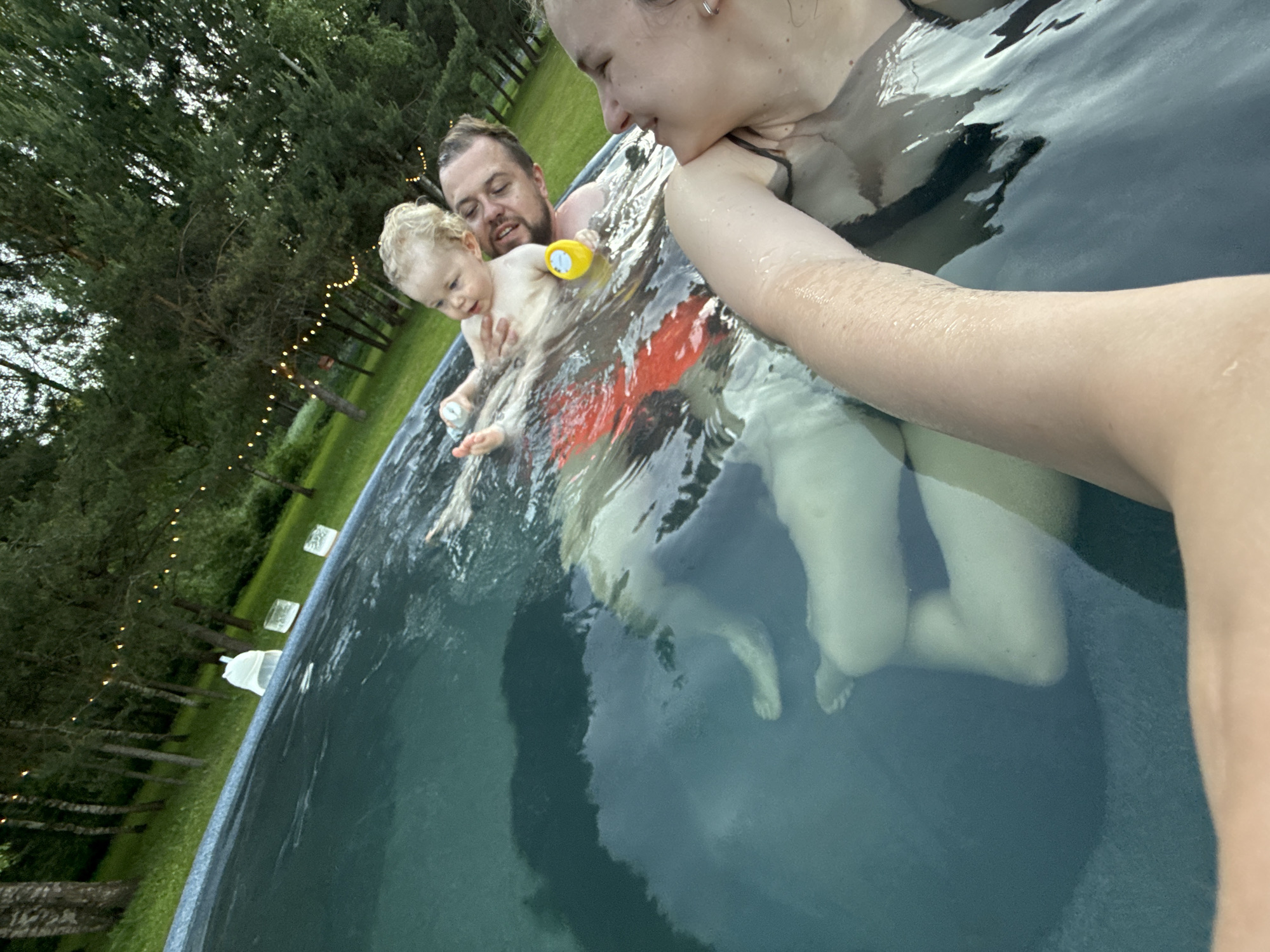 In an outdoor swimming pool in Копнино, Russia, a family is observed. In the immediate foreground, a female subject's right arm, shoulder, and the left side of her head are visible as she captures a selfie. Her head is turned slightly to the right, exhibiting a subtle smile. She wears a dark-colored swimsuit top and a small silver earring in her left ear. Further into the pool, a male adult with short dark hair and a beard is partially submerged, smiling while looking towards a young child he is holding. The child, with light, curly hair, looks slightly to their left, holding a small white object in their right hand. A yellow plastic toy floats near the child's left hand. Submerged below the water surface, red fabric is visible, consistent with swim shorts worn by the man or another person nearby. Along the far edge of the pool, two translucent rectangular objects and a white, soft item are positioned. The pool features a blue-patterned liner. Beyond the pool edge, a green lawn transitions into a dense natural environment comprising various trees, including lighter-barked deciduous trees and darker evergreen conifers. String lights with warm illumination are strung between the trees, indicating the time is likely dusk or evening.