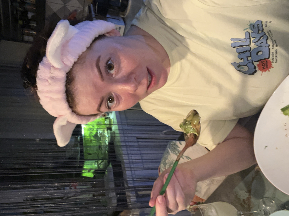 A woman wearing a pale yellow T-shirt with "Chill Days" printed on it and a pink bunny ear headband is holding a spoon of green food. She is looking at the camera with a slight smile. The background includes a view of greenery, a metal fence, and a white plate with food remnants.