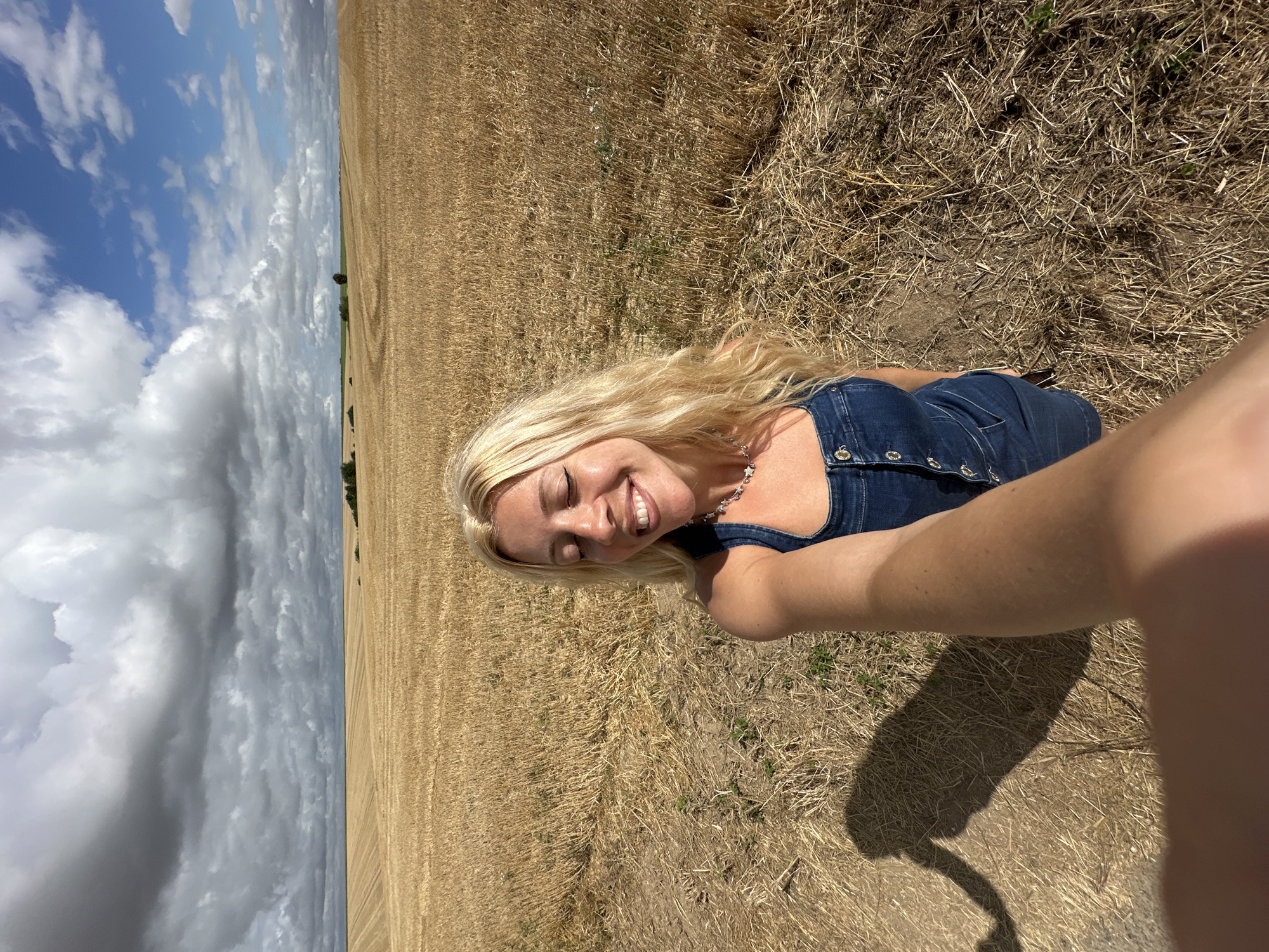A young woman with blonde hair and closed eyes, smiling, takes a selfie in a dry field. She is wearing a denim jumpsuit and a necklace. The background is a vast, dry field with scattered trees and a cloudy sky. The image is taken from a high angle, with the woman in the foreground and the field stretching to the horizon. There is no indication of the city of Royon, France in the image.