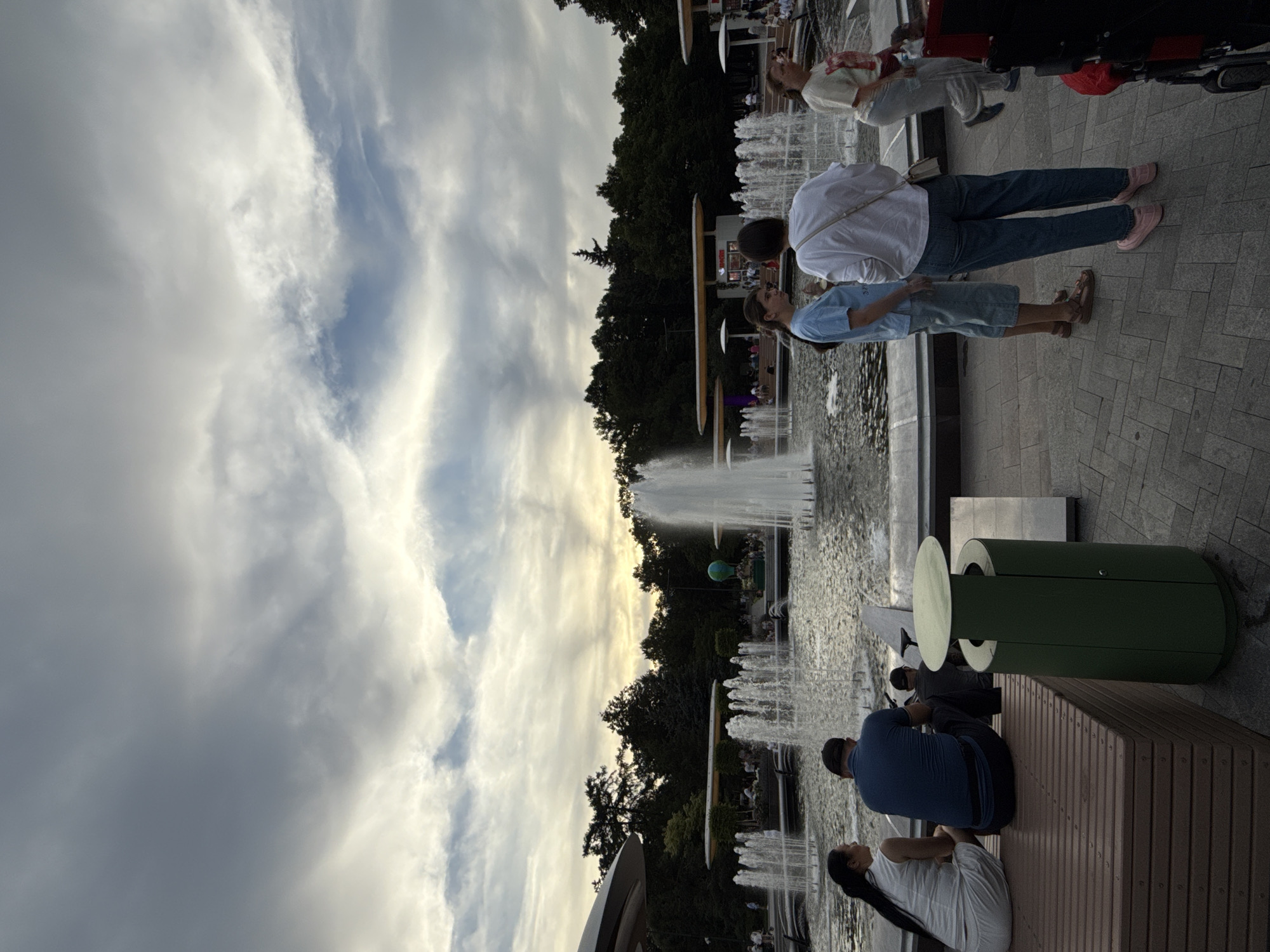 This is a medium-shot, eye-level, outdoor photograph taken at the VDNKh park in Moscow, Russia. The image captures a fountain area with several people present.
In the foreground, a man in a blue t-shirt and a woman in a white top are sitting on a wooden bench, facing away from the camera. To their right, a green trash receptacle stands on the paved ground.
In the mid-ground, four individuals are standing and interacting. Two young women in casual attire are conversing, one in a blue t-shirt and shorts, the other in a white long-sleeved shirt and jeans. Further to the right, a woman in a light-colored t-shirt and pants is walking, and another person is partially visible in the far right of the frame.
The background features a large, circular fountain with multiple jets of water spraying upwards. Beyond the fountain, lush green trees form a dense line, with some park structures and a hot air balloon sculpture visible amongst them. The sky above is cloudy, with patches of light breaking through, suggesting it is either early morning or late afternoon. The overall atmosphere appears relaxed and public.