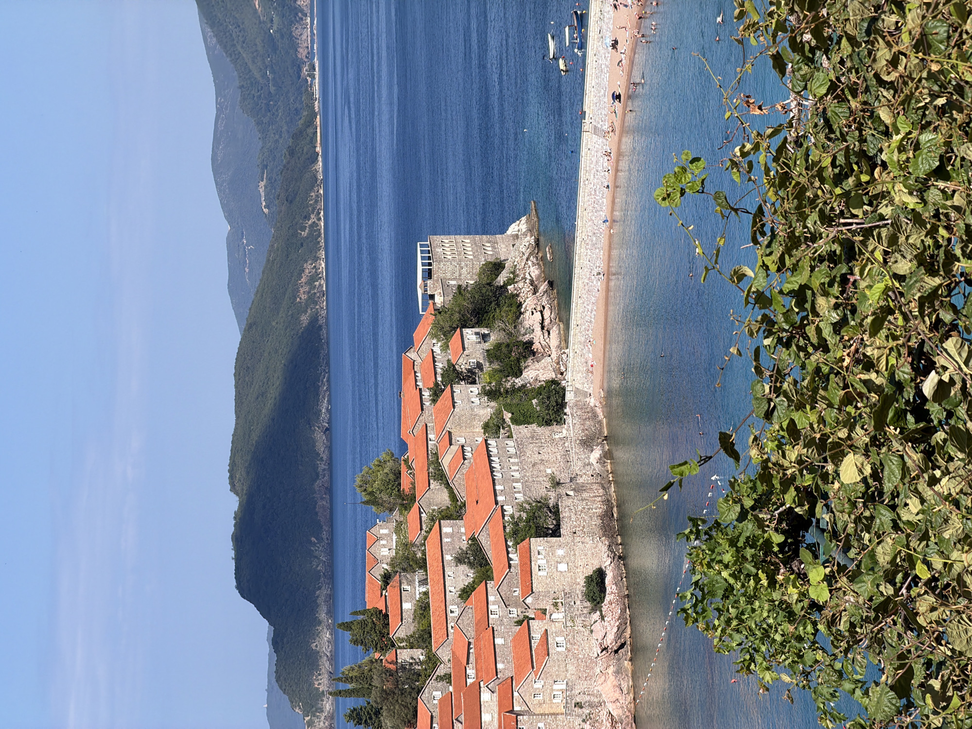 The image shows the Sveti Stefan islet in Montenegro, a small fortified island village connected to the mainland by a causeway. The islet is densely packed with stone houses featuring distinctive red-tiled roofs. The buildings appear to be a mix of residential and perhaps hotel accommodations, with some showcasing multiple stories and stone walls.
The causeway leads to a sandy beach where numerous people are visible enjoying the sunny weather. Some are sunbathing, while others are in the water, swimming or wading. Several small boats are moored in the clear blue sea surrounding the island and beach.
In the background, rolling green hills and mountains rise from the coastline, with the sea stretching out to the horizon under a clear blue sky. The foreground is partially obscured by lush green foliage, adding depth to the scene. The overall impression is one of a picturesque coastal resort bathed in sunlight.