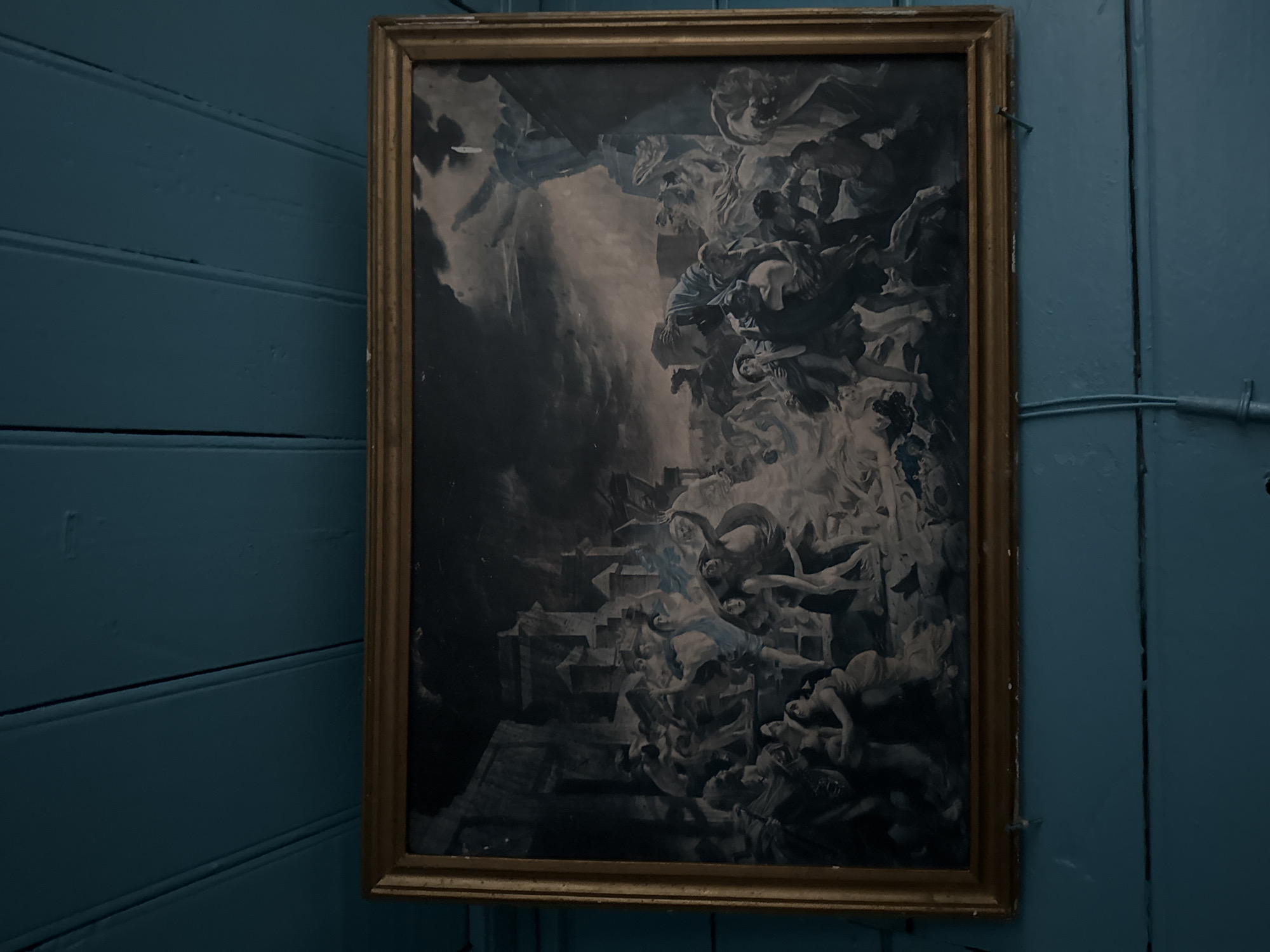 The image shows a framed painting with a monochromatic, dramatic scene. The painting depicts a chaotic and violent event with numerous nude or partially draped figures in distress. The figures appear to be fleeing or struggling amidst collapsing structures and swirling clouds, suggesting a catastrophe such as an earthquake, fire, or divine judgment. The style of the painting suggests it is a historical or biblical depiction, possibly a scene from mythology or a significant event. The background is dark and stormy, with hints of architectural elements. The painting is hung on a teal-colored wooden paneled wall. There is no identifiable location of Sosnovskoye, Russia present in the image or its context.