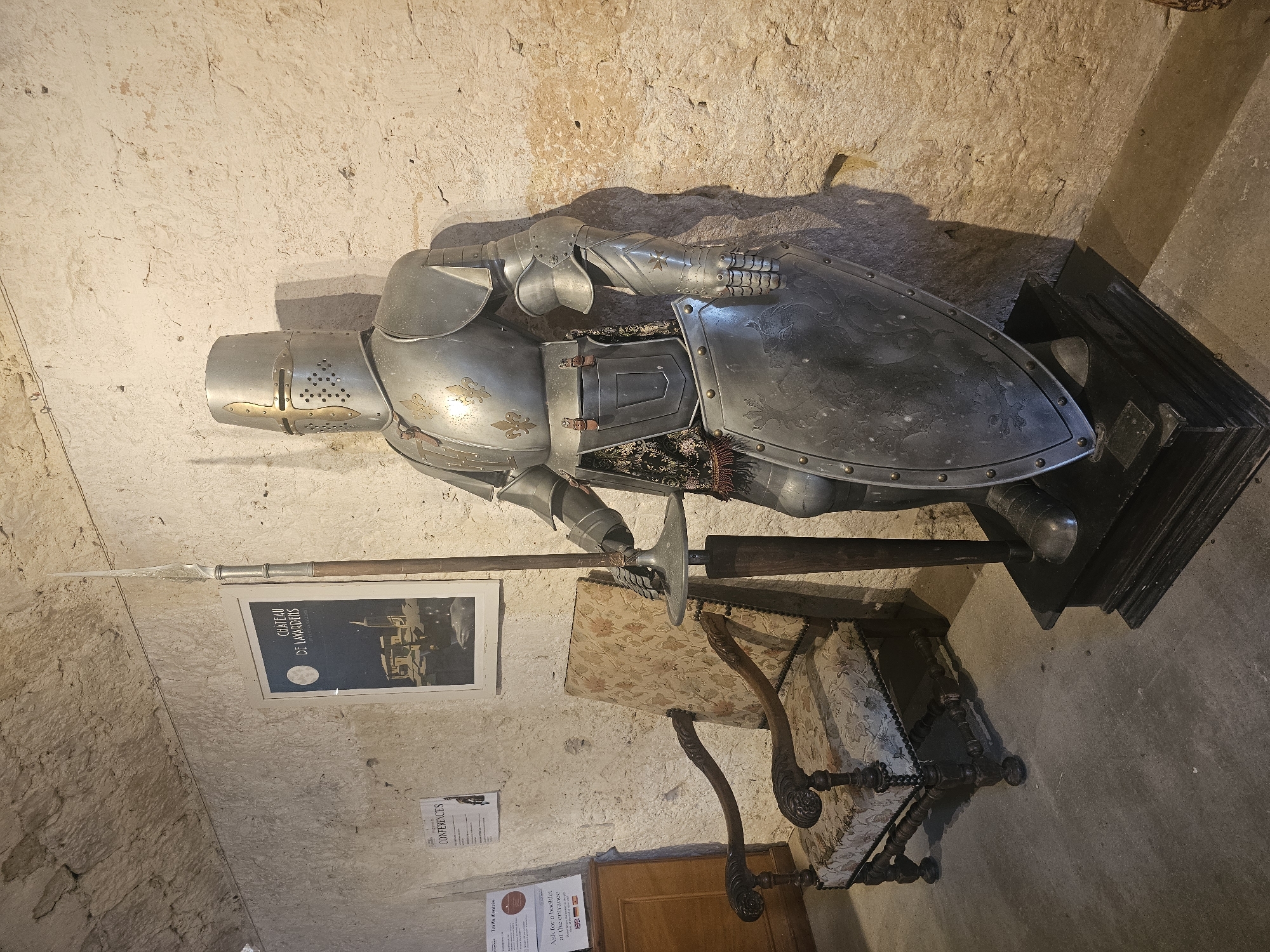The image shows a full suit of medieval armor displayed on a black platform in a dimly lit, stone-walled room. The armor is made of polished metal, with decorative gold fleur-de-lis emblems on the chest and shoulders. It includes a helmet with a visor, pauldrons, gauntlets, a breastplate, and greaves. A large, ornate shield with embossed patterns rests in front of the armor. A long spear with a pointed tip is held upright by the armor. To the left of the armor, there is an antique wooden armchair upholstered in a floral fabric. Behind the armor and chair, a poster is affixed to the wall, featuring the text "CHÂTEAU DE LAVARDENS" and an illustration of a castle under a full moon, indicating the location as the Château de Lavardens in Lavardens, France. To the far left, part of a wooden cabinet and two informational signs are visible. The floor is concrete.