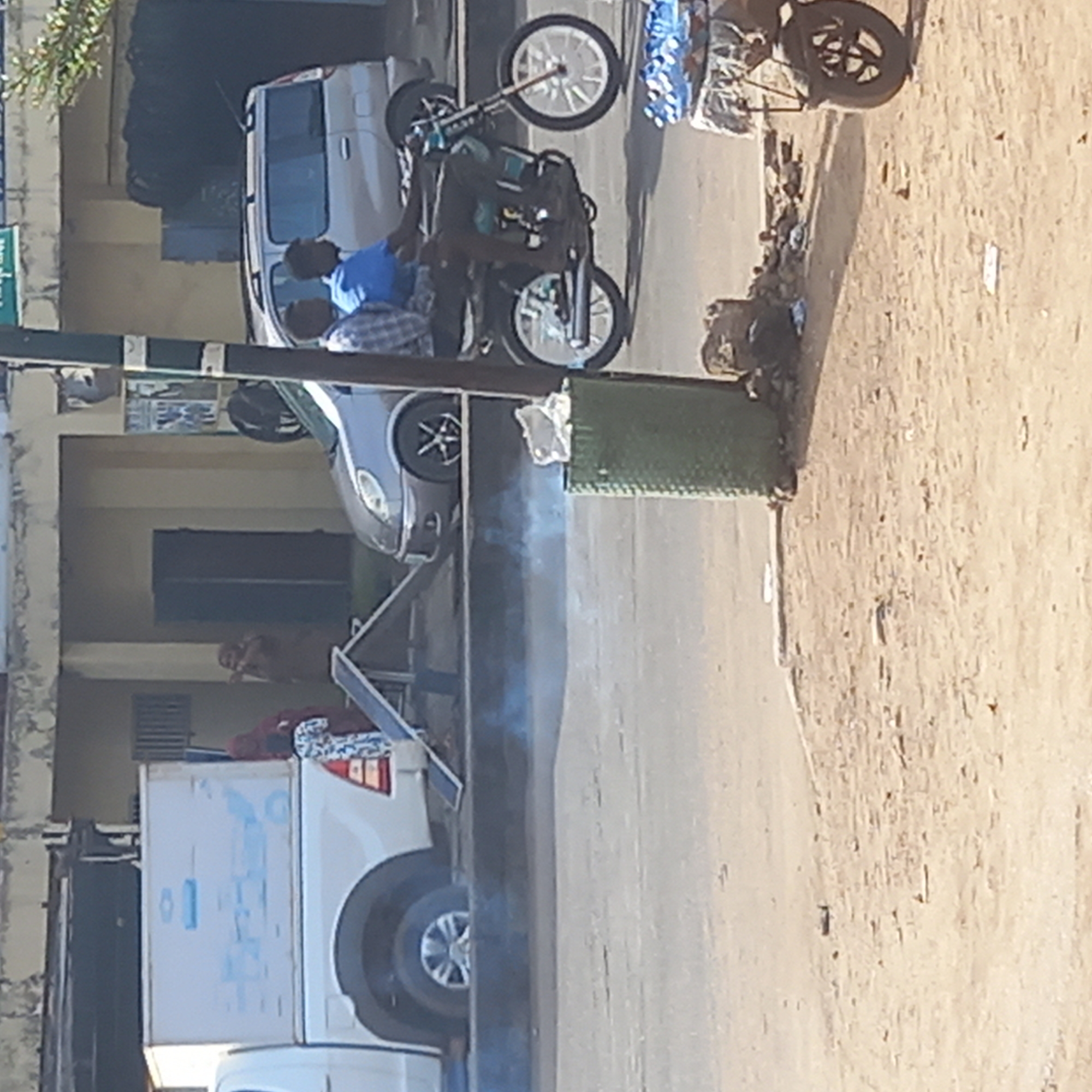 An outdoor scene in Dadi Riba, Nigeria, depicts a street with several vehicles and people under bright daylight. The ground is a mix of paved road and dusty earth.
In the foreground, two men are actively engaged with a black motorcycle that features prominent silver-colored spoked wheels. The motorcycle appears to be propped up, with one of its wheels elevated, suggesting maintenance, repair, or inspection. One man wears a blue shirt, and the other a plaid shirt.
Behind the motorcycle, a light-colored minivan or SUV is parked. Further down the street, on the right side, a white cargo van with blue graphics and indecipherable text on its side is visible. Another light-colored car is partially obscured behind a green, textured barrier or structure.
To the far right, a small, two-wheeled cart is parked near a pile of debris. A faint plume of white smoke rises from the ground near the green barrier and the white van. Buildings with visible windows and entrances line the left side of the street, with a partially visible person inside one entrance further back. A dark pole, likely a lamppost or signpost with an unreadable green sign, stands on the left. The clear daylight indicates it is likely daytime, with no signs of adverse weather.