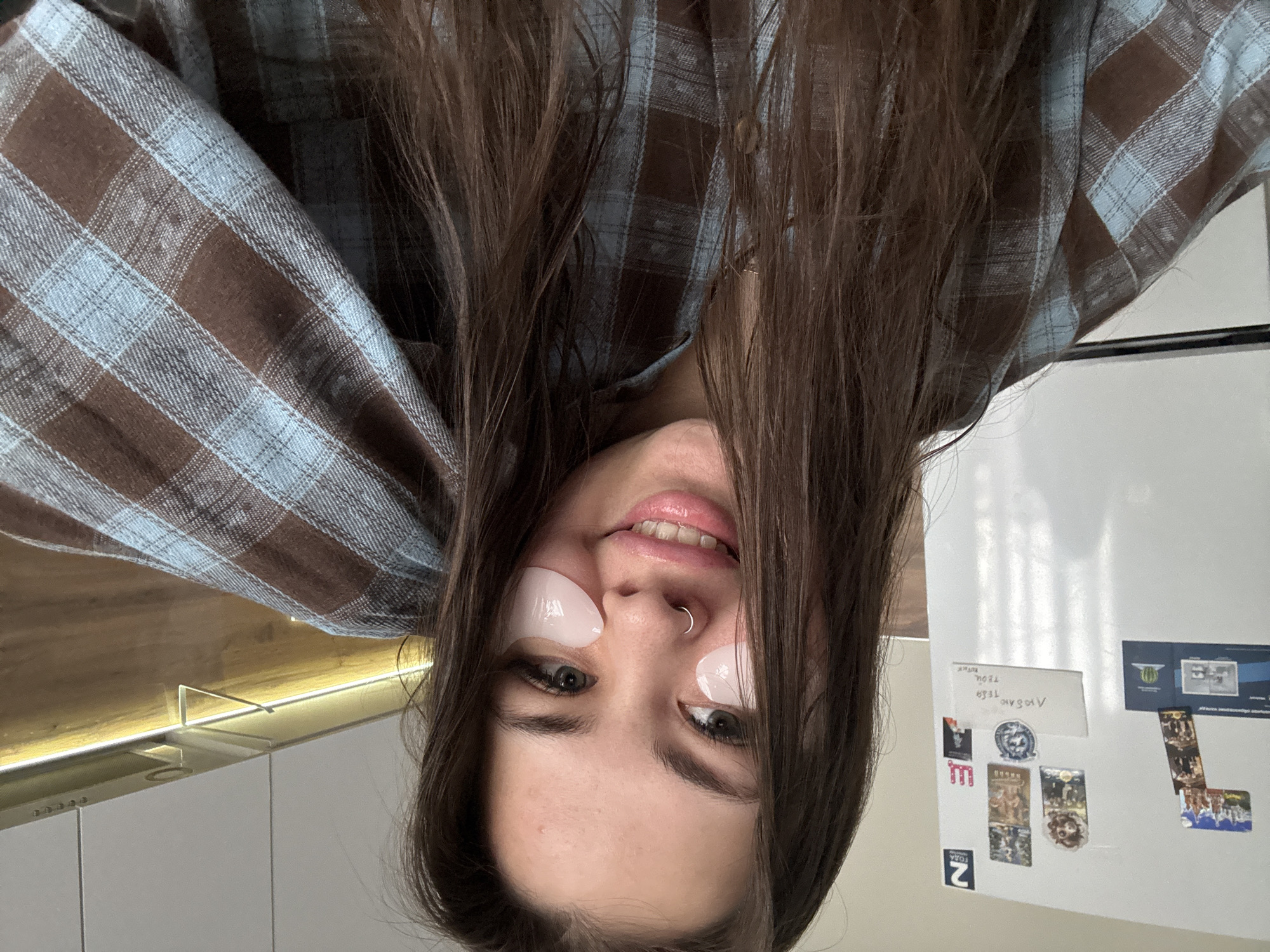 A casual self-portrait captures a young woman from a low angle, with her face appearing upside down in the frame. She has long, dark brown hair flowing downwards and is wearing clear, gel-like under-eye patches on her cheeks below each eye. A silver nose ring adorns her left nostril, and her lips are slightly parted in a relaxed, subtle smile, showing a hint of her teeth. She is dressed in a brown and light blue plaid flannel shirt.
The background reveals a domestic interior, likely a kitchen in Bugry, Russia. To her right (image left), a wooden surface is visible, illuminated by a strip of warm yellow LED lighting, beneath which are white kitchen cabinets. To her left (image right), a white refrigerator is covered with various magnets, including small pictures, postcards, and at least two handwritten notes. One note, appearing upside down from the viewer's perspective, features text that is difficult to decipher due to orientation and handwriting; however, when rotated and inverted, it could be interpreted as Cyrillic characters, possibly 'АНОХОV' (Anokhov, a Russian surname) and 'ТБЛЦ'. Another smaller label reads '2 FOKA' or '2 FODA'. The overall scene suggests a relaxed, casual moment at home.