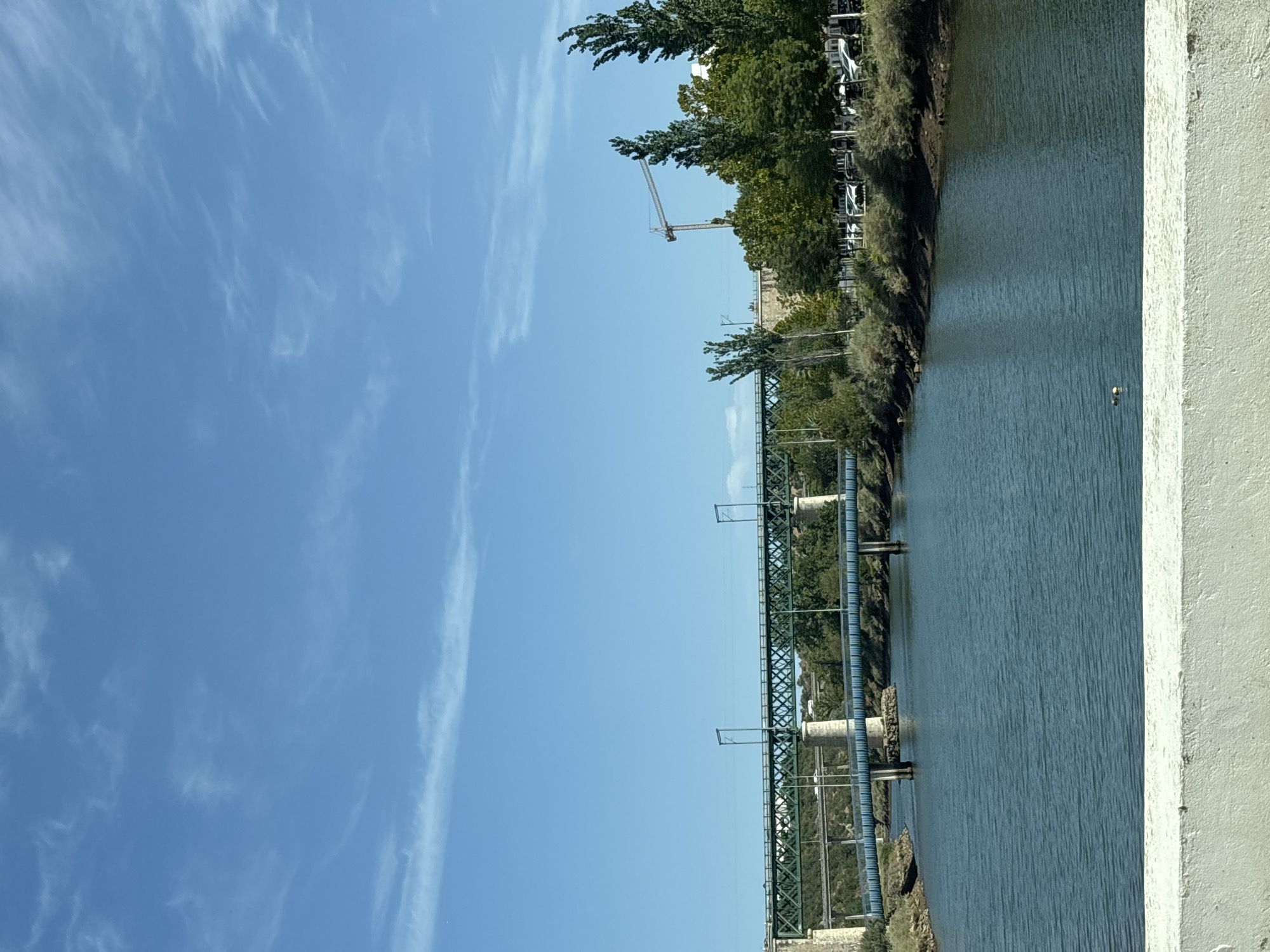 The image depicts a wide, calm river in Tavira, Portugal, under a bright blue sky with wispy white clouds. Two bridges span the river. The foreground bridge is a dark blue, modern-looking structure. Further back, a larger, green, lattice-style railway bridge is supported by stone piers. Trees line the riverbanks, with a construction crane visible in the background behind a cluster of tall, slender trees and a line of parked cars. The foreground of the image is dominated by a white concrete barrier. The scene appears to be a daytime shot, likely taken from a slightly elevated position, possibly from another bridge or embankment.