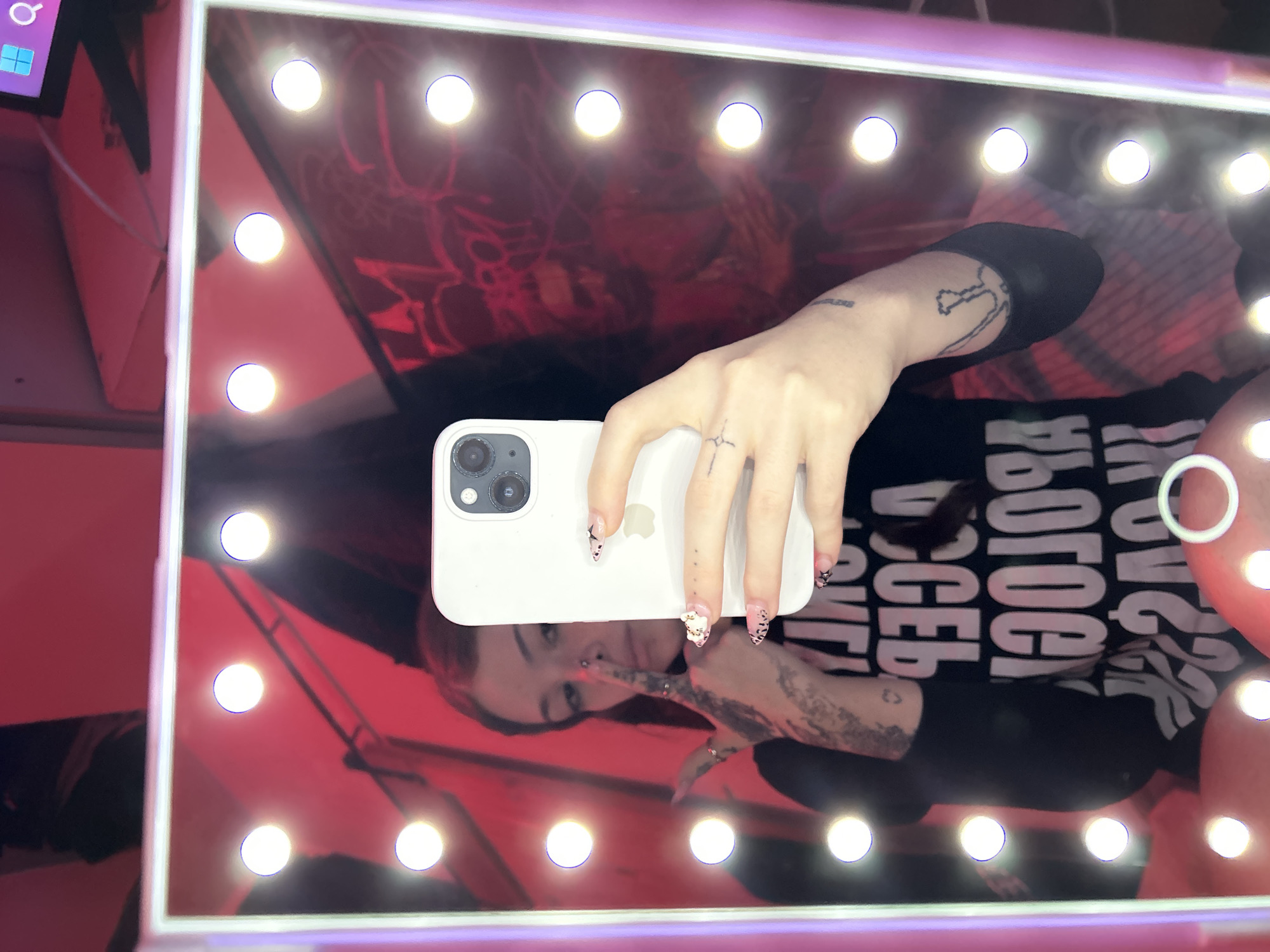 A person is reflected in a mirror with light bulbs lining its perimeter. The person is holding a white smartphone with three rear cameras in their right hand. The fingernails on their right hand have decorative designs. The person's right arm has tattoos. The person is wearing a black shirt with white writing on it. The reflection includes other objects in the background with a red hue. A portion of a monitor is visible in the upper left. The location is a dressing room. There is no information in the image that would describe the city of Buenos Aires, Argentina.