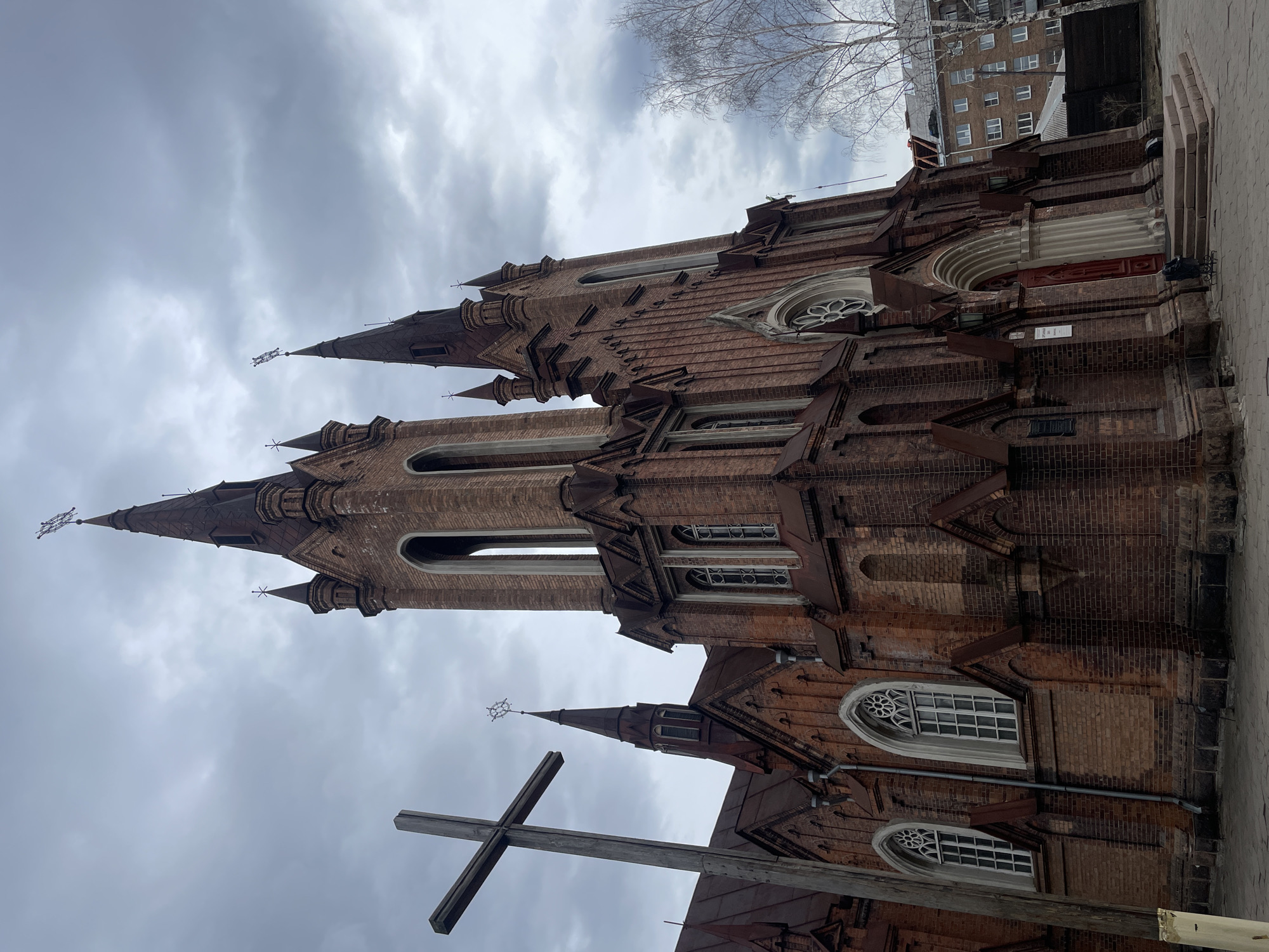 The image depicts the Roman Catholic Church of the Transfiguration in Krasnoyarsk, Russia. The church is built of red brick in a Gothic Revival style, featuring tall spires, arched windows with decorative tracery, and buttresses. A large wooden cross stands in the foreground. The sky is overcast, and bare tree branches are visible in the background. A residential building is visible to the right of the church. The photo is taken from a low angle, looking upwards at the church.