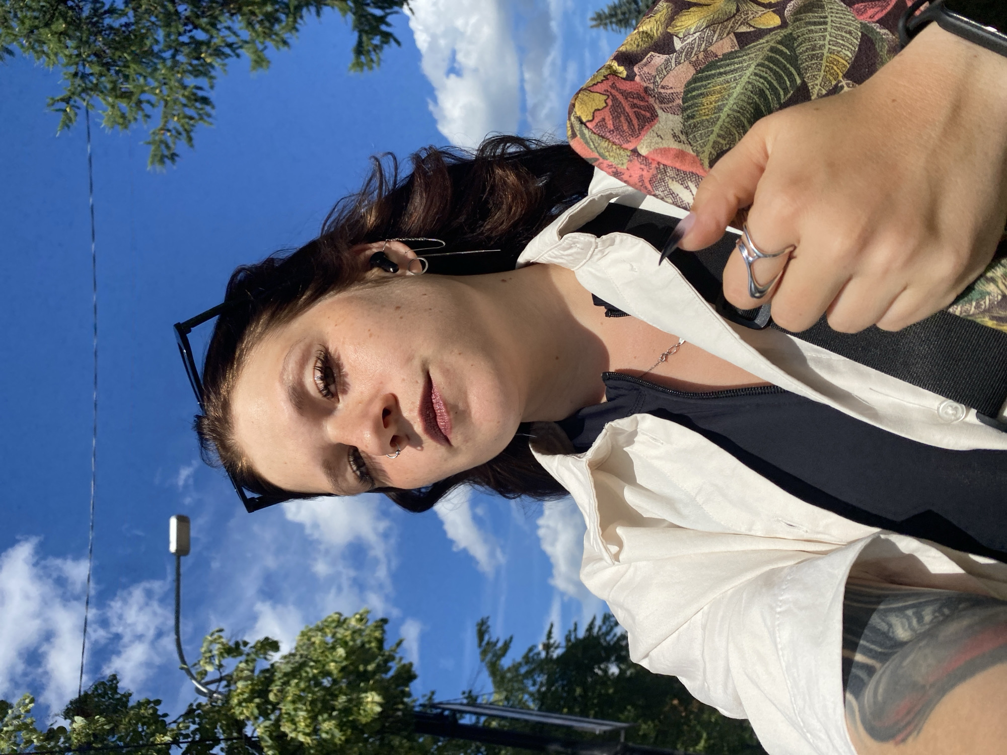 A young woman with long, dark, wavy hair is taking a selfie outdoors. She is wearing a black top under an unbuttoned white shirt. She has a nose ring, earrings, and rings on her fingers. She is holding a floral patterned bag on her right side. Her left arm, visible in the lower left corner of the image, has a tattoo. The background shows a bright blue sky with scattered clouds, trees, and a street lamp. The scene appears to be a sunny day in a city, potentially Chaikovsky, Russia, given the context.