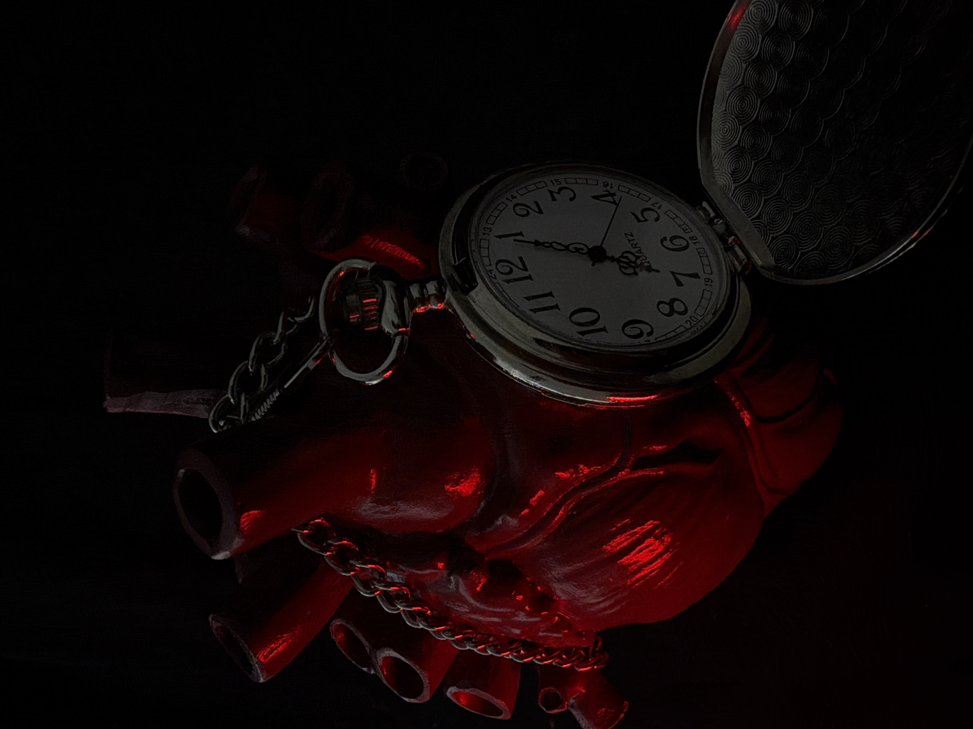 A close-up, low-angle shot features a red, anatomical heart sculpture against a black background. A silver pocket watch with a chain is draped over the sculpture's left side, open to reveal its face. The watch face displays the time as approximately 12:05. The heart sculpture has a rough, textured surface and appears to be lit from the left, casting strong red highlights and deep shadows. The overall lighting is dim and dramatic. The background is entirely black, with no discernible details of any location.